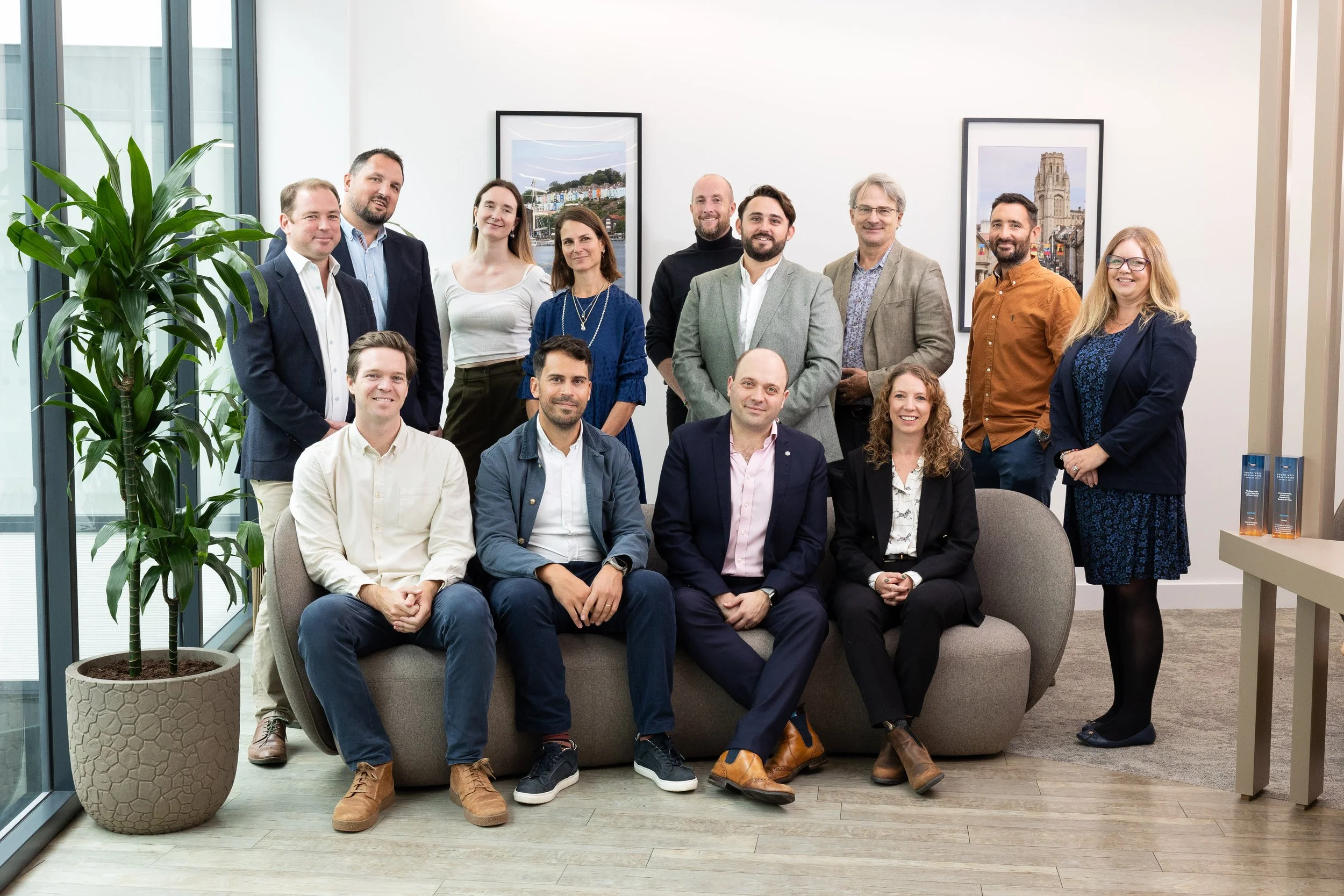 A group of 15 diverse professionals in business attire posing in office with framed photos and a large potted plant.