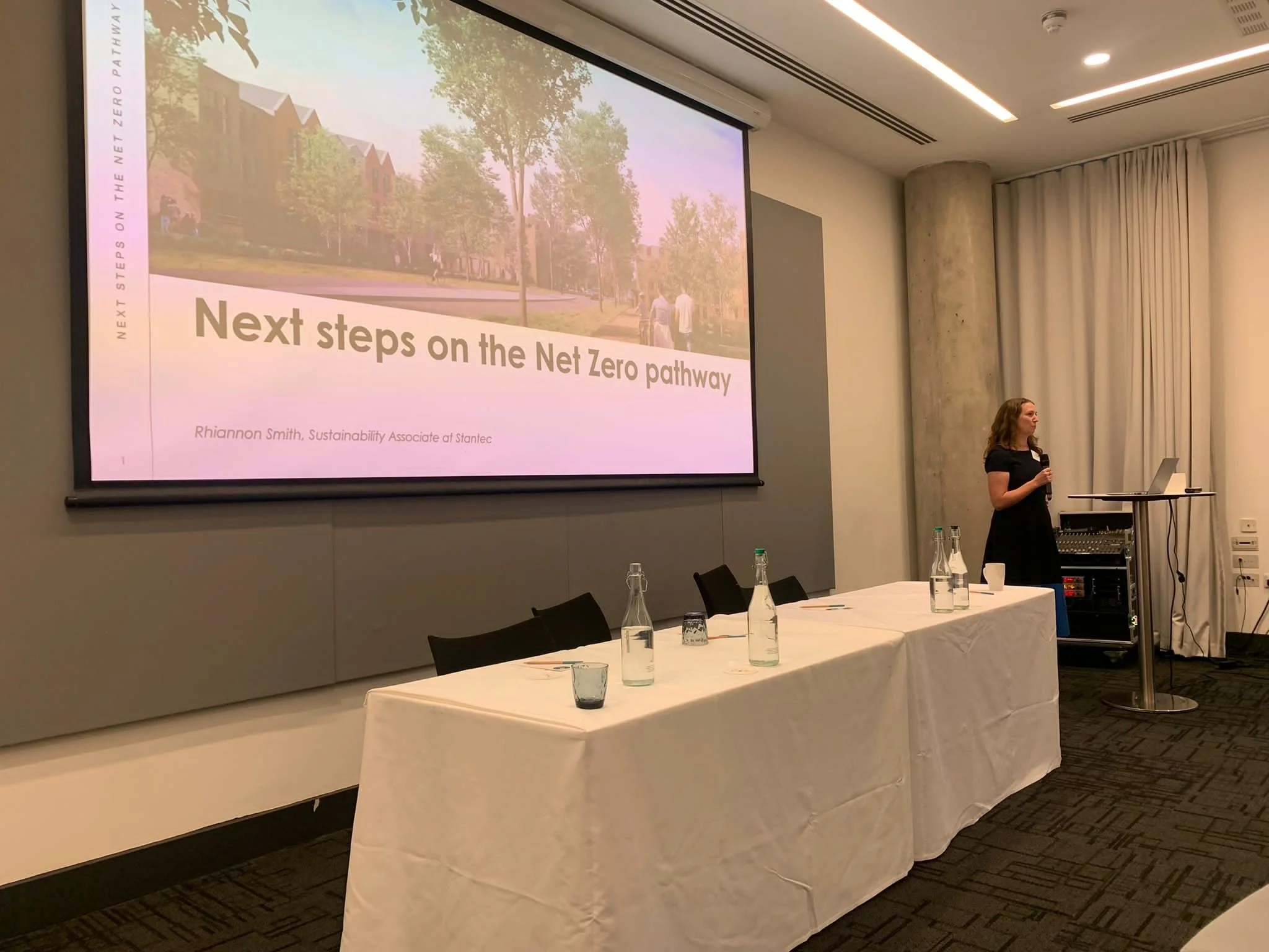 A photo of Rhiannon Smith standing on stage giving a presentation titled 'Next steps on the Net Zero pathway' in a conference room with a large screen, tables with water bottles, and curtains.