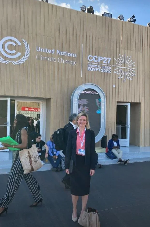 A photo of Lucy Wood in business attire standing outside a conference center with signage for the United Nations Climate Change Conference (COP27) held in Egypt in 2022, with people sitting and walking nearby.