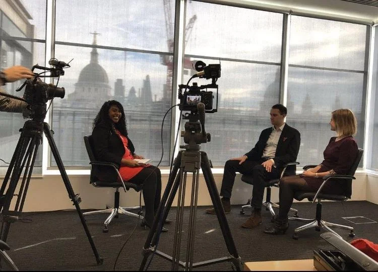 A video recording session with three individuals, two women and one man, seated in a brightly lit room with large windows and city skyline in the background, while a camera on a tripod records them and a person operates another camera.
