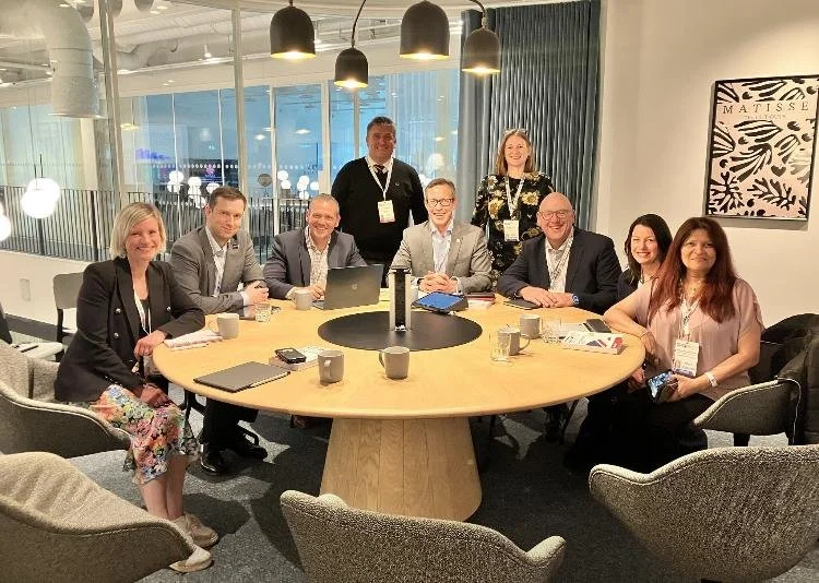 Group of nine professionally dressed people sitting and standing around a round conference table in a modern office or hotel meeting room, with laptops, notebooks, mugs, and conference badges, during a business meeting or conference.