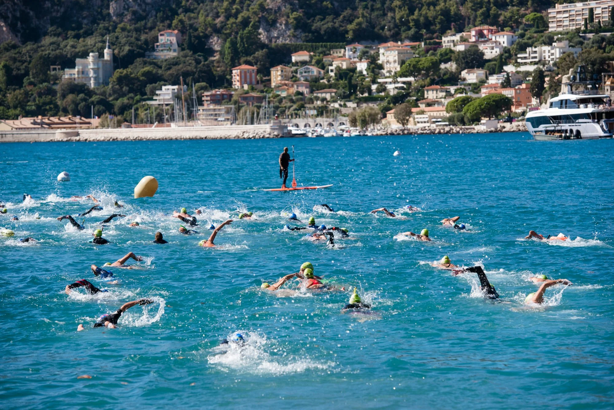 Natation de triathlon avec plusieurs nageurs portant des bonnets colorés dans une mer bleue, avec un entraîneur sur une planche à proximité, et une ville avec des maisons et des bateaux dans le fond.