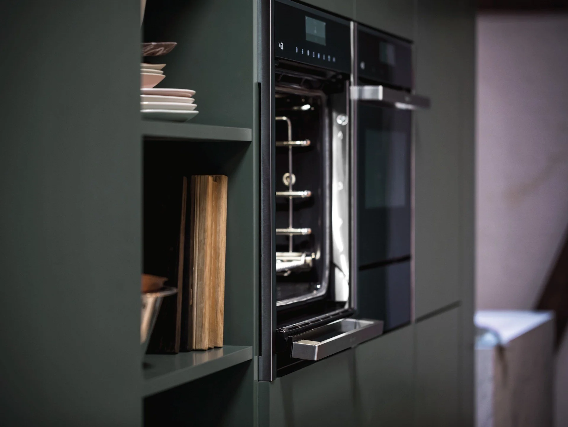 A kitchen with a built-in oven, shelves with plates, and a cutting board.