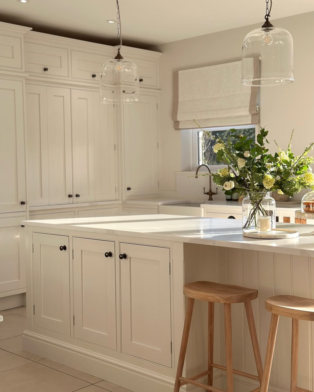 Kitchen with white cabinets, island, window with Roman shade, hanging glass light fixtures, and a vase with flowers on the counter