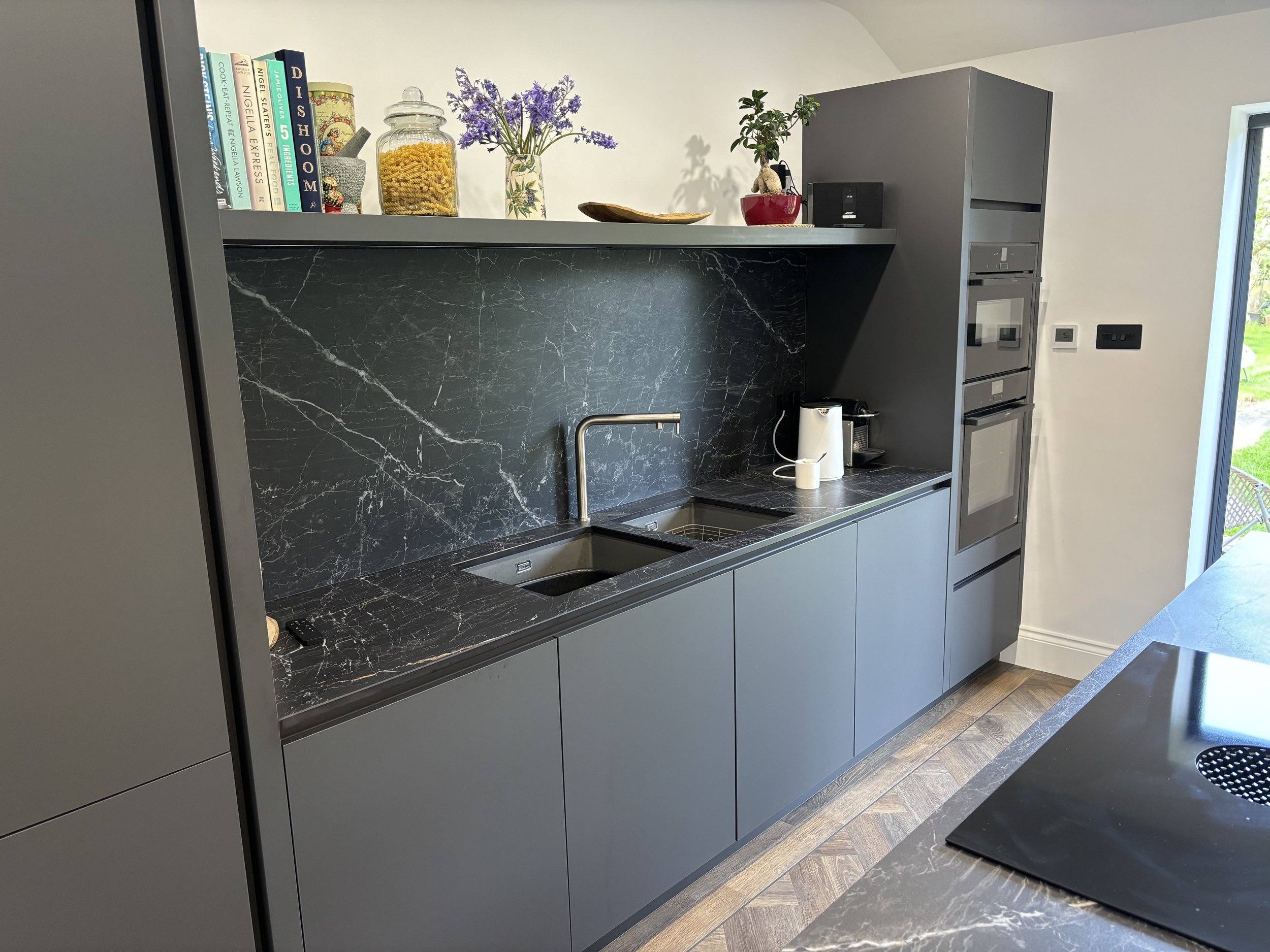 Modern kitchen with black marble countertops and backsplash, gray cabinets, and built-in oven. Upper shelf with books, flowers, and decorative items. Small appliances on the counter, including a kettle and coffee machine. Part of a window showing outdoor greenery.