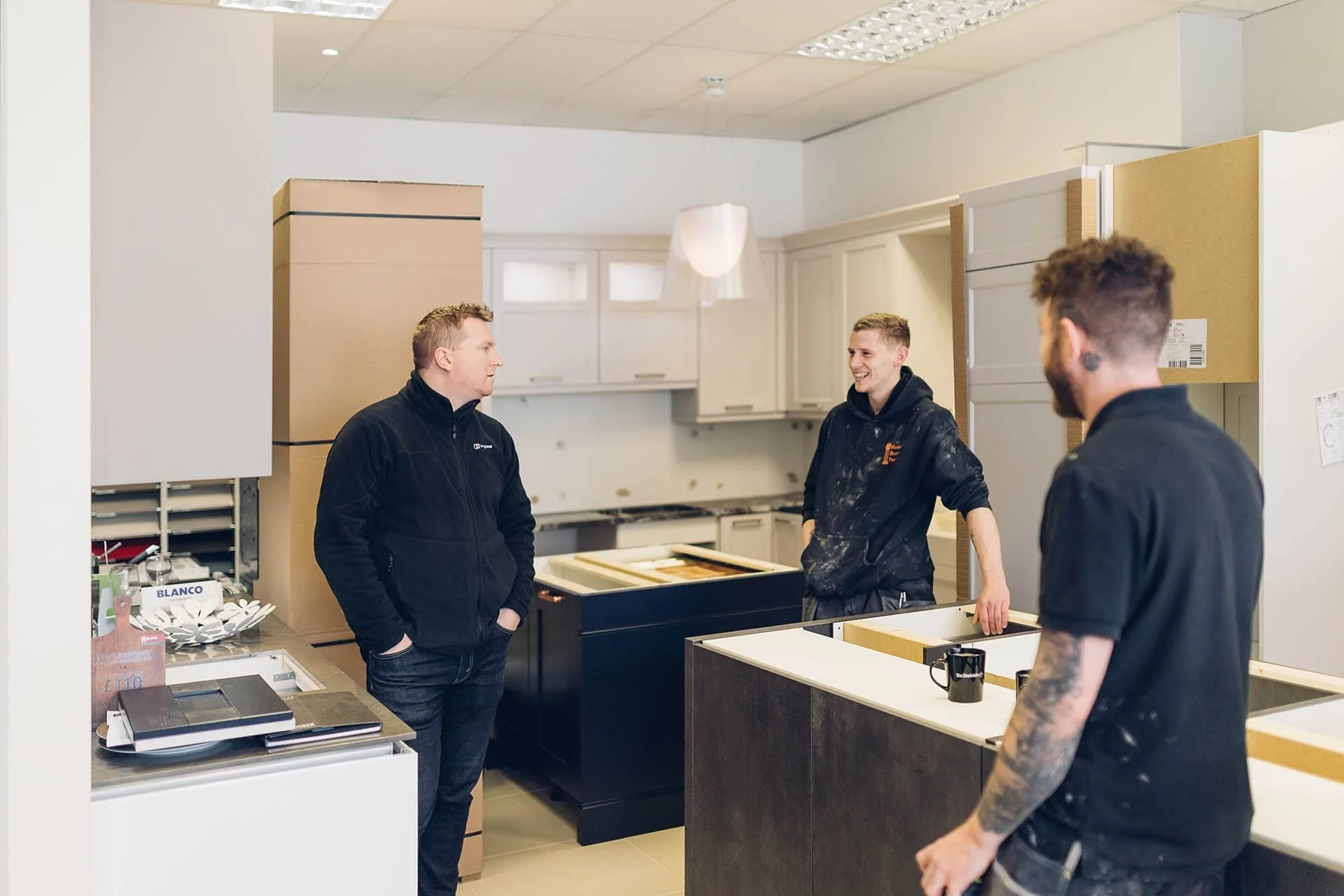 Three men are standing and talking in a kitchen showroom, with cabinets and countertops, one man is wearing a black jacket, and the other two are in black hoodies, one of whom has tattoos on his arm.