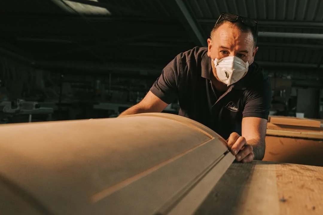 A man wearing a face mask and glasses on his head inspects a wooden object in a workshop.