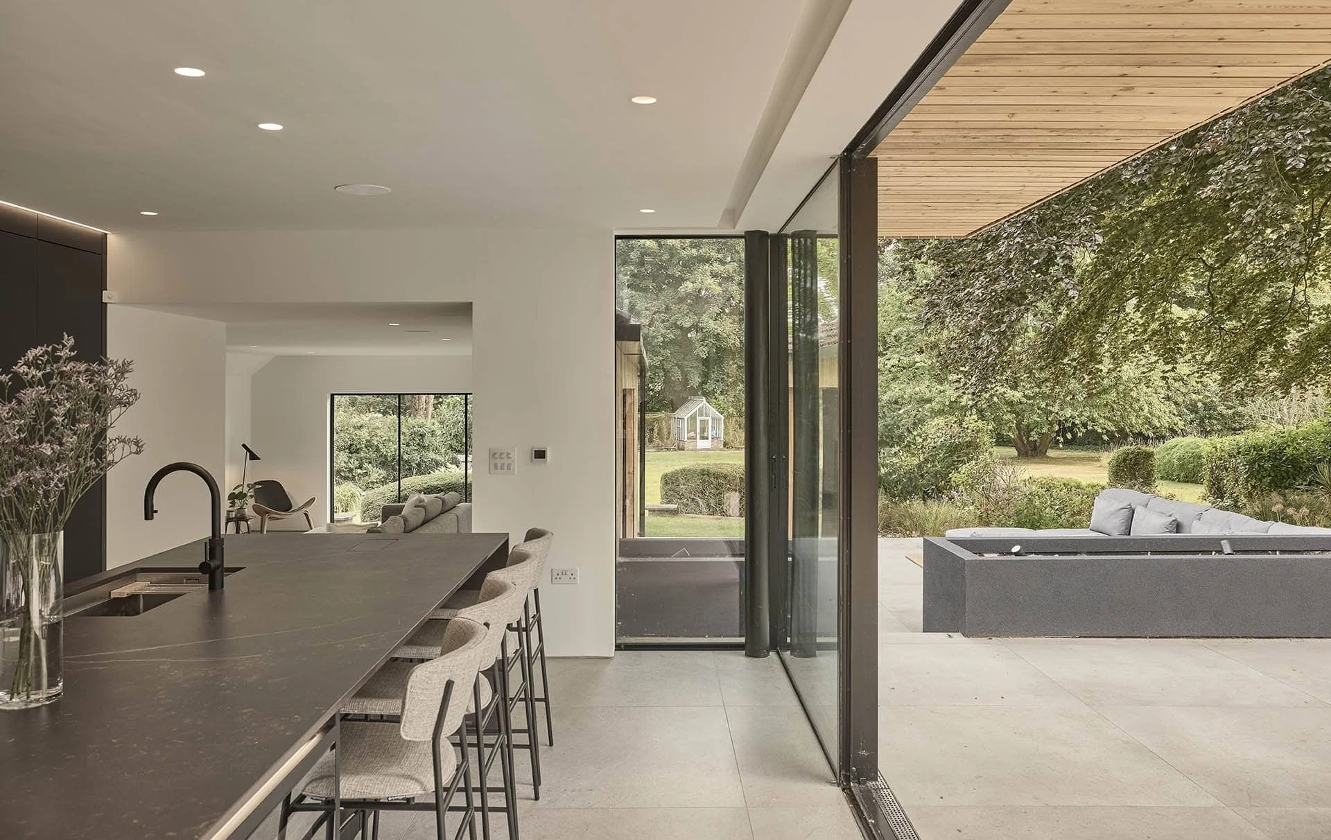 Modern open-plan kitchen and living room with large sliding glass door opening to outdoor patio and garden, featuring minimalist decor, neutral color palette, and outdoor seating.