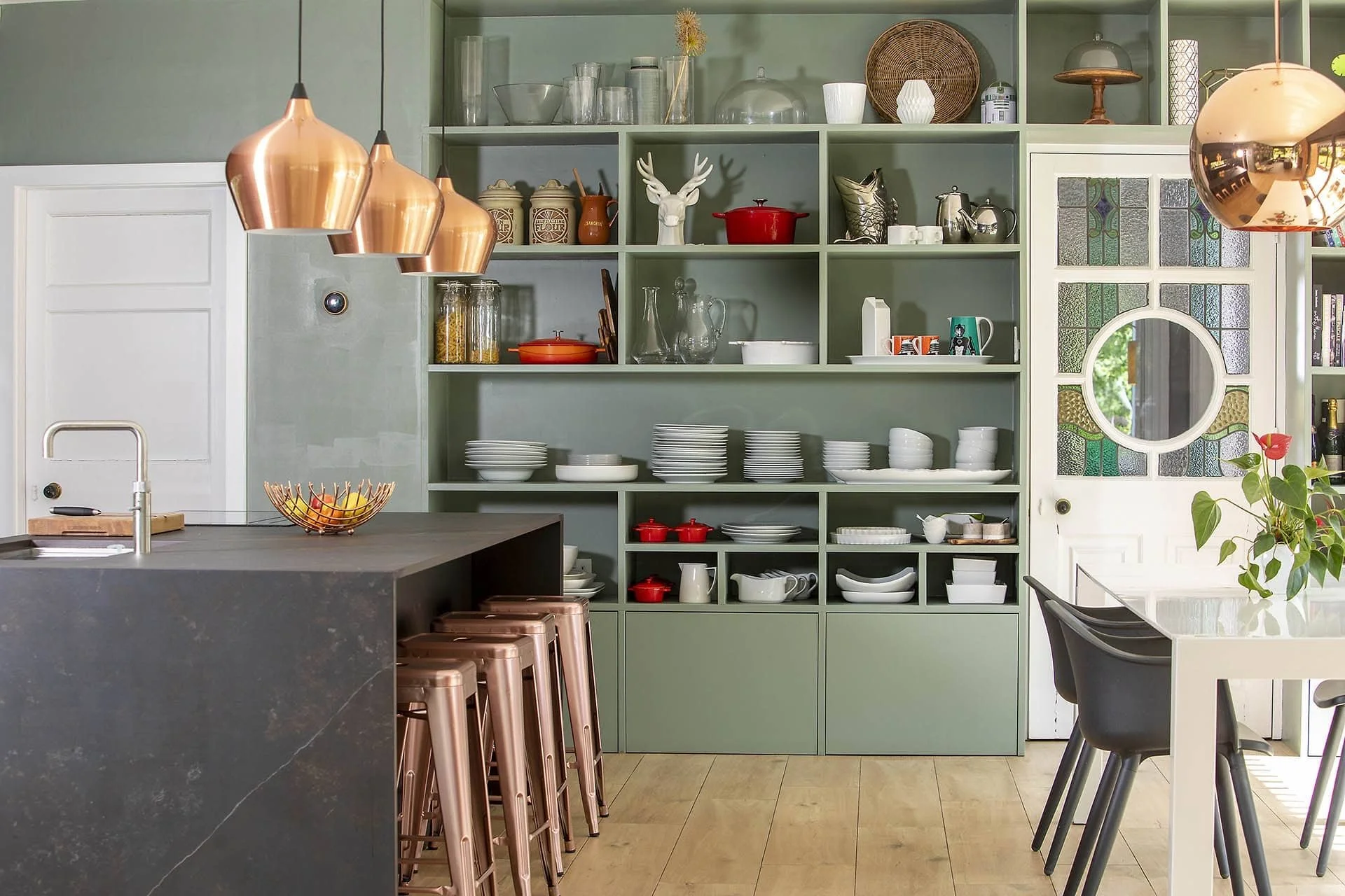 Open green kitchen shelving with dishware, decorative objects, and glass jars, adjacent to a white door with stained glass accents, and a kitchen island with a dark stone countertop and copper pendant lights.