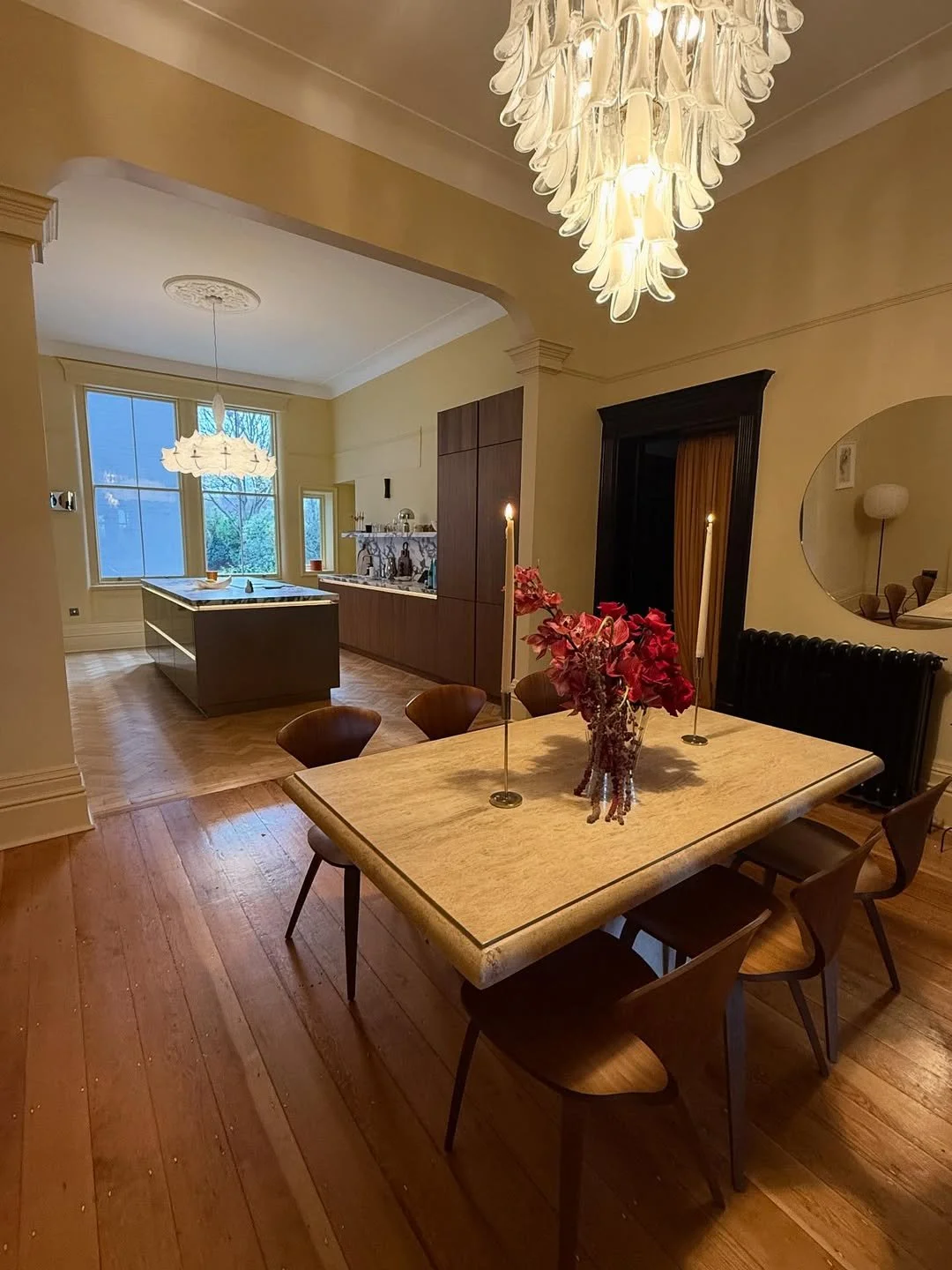 A dining room with a wooden table and six chairs, floral centerpiece, and two lit candles. The room has a chandelier, a window, and a kitchen area with an island in the background.