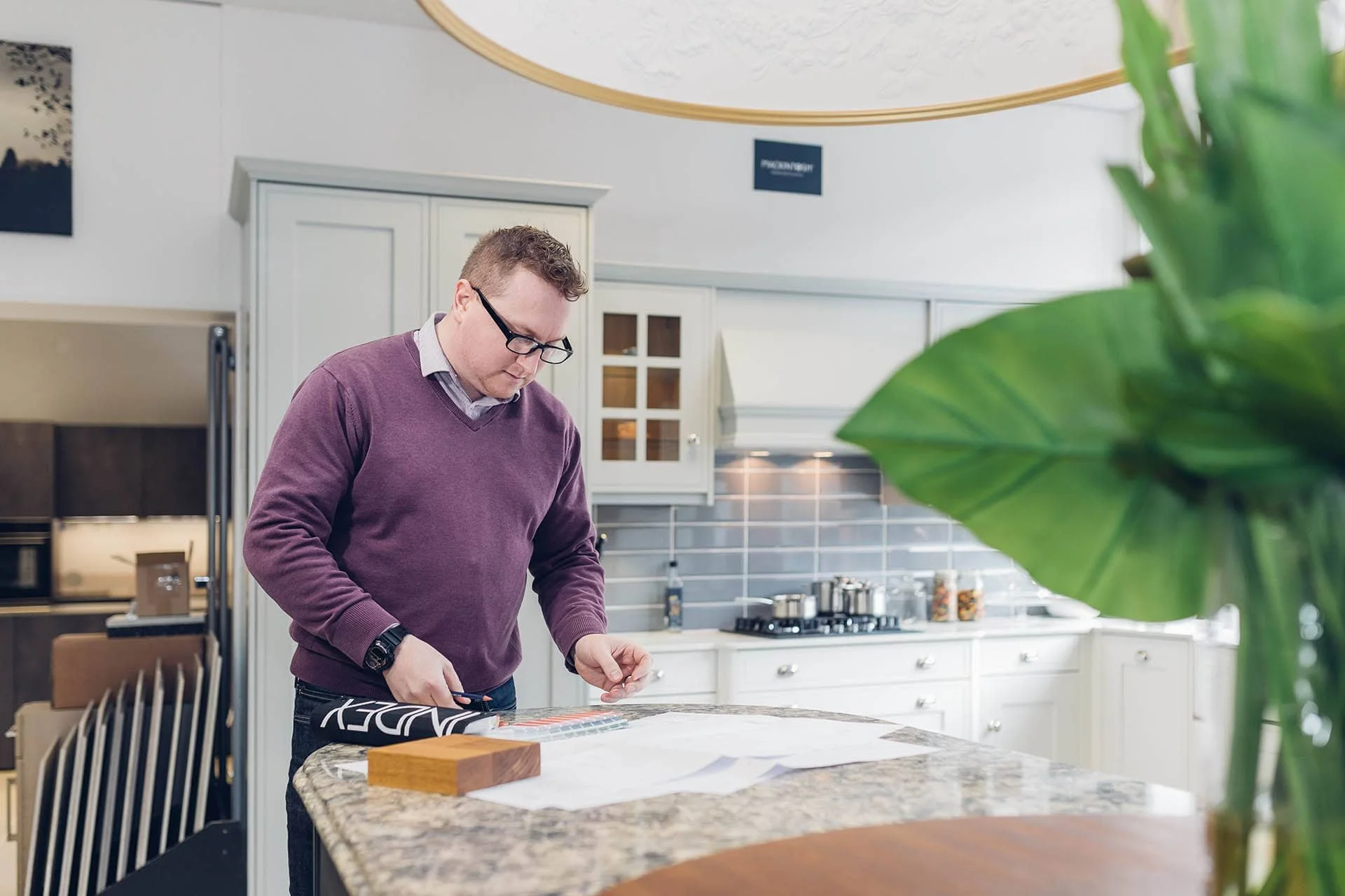 A man in a purple sweater and glasses standing at a kitchen counter, cutting a piece of fabric with scissors.