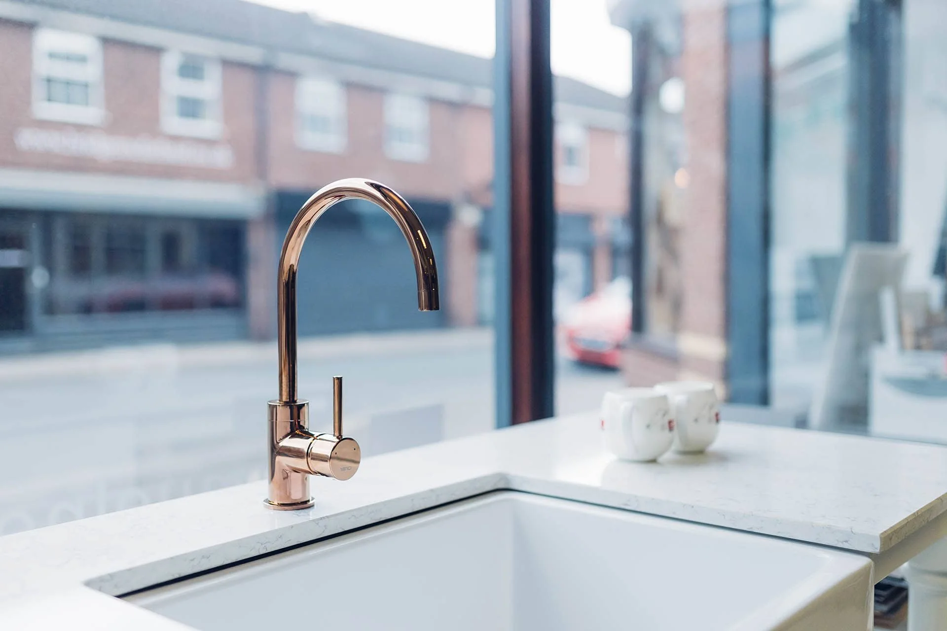 Modern kitchen sink with a rose gold faucet and white countertop, with three white cups on the right, near large windows overlooking a city street.