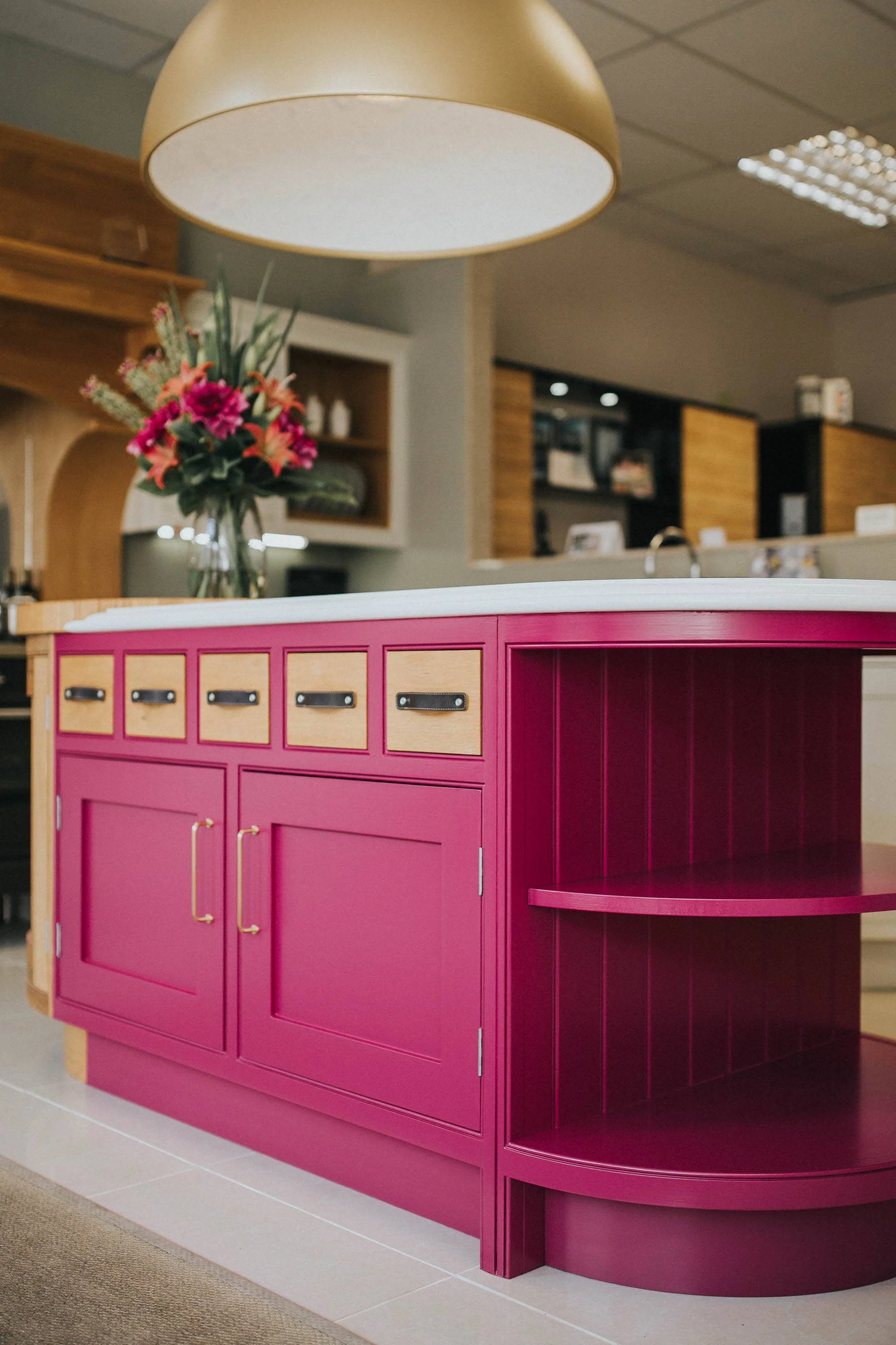 A pink and wood-colored cabinet with a white countertop and drawers, located in a room with a hanging lamp and a vase with flowers in the background.
