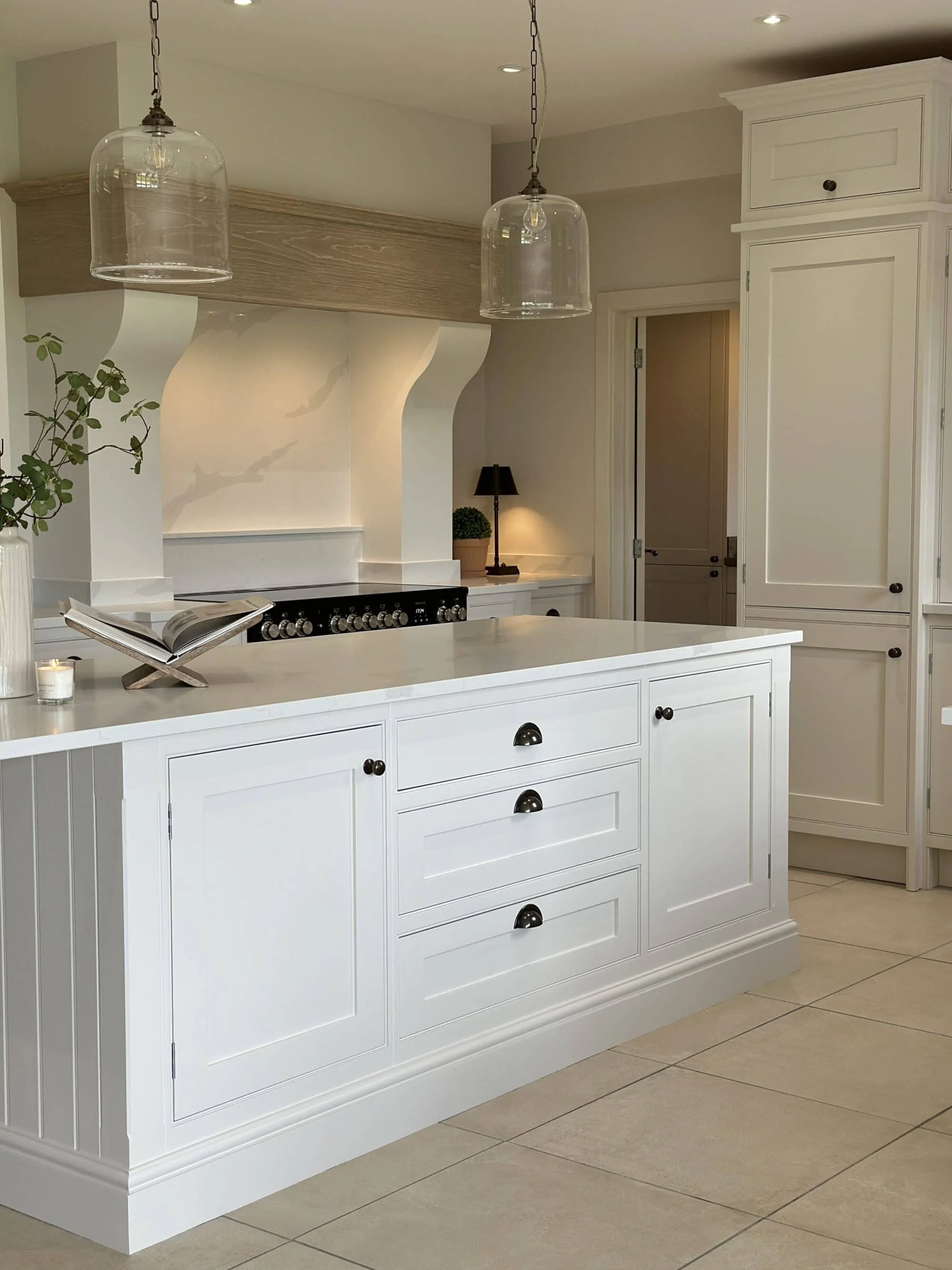 A modern kitchen with white cabinetry and a white kitchen island, black cabinet handles, pendant lights, and a stove with a vent hood. There are decorative items on the island and in the background, including a potted plant, a small lamp, and a book. The floor is tiled, and the overall color scheme is neutral.