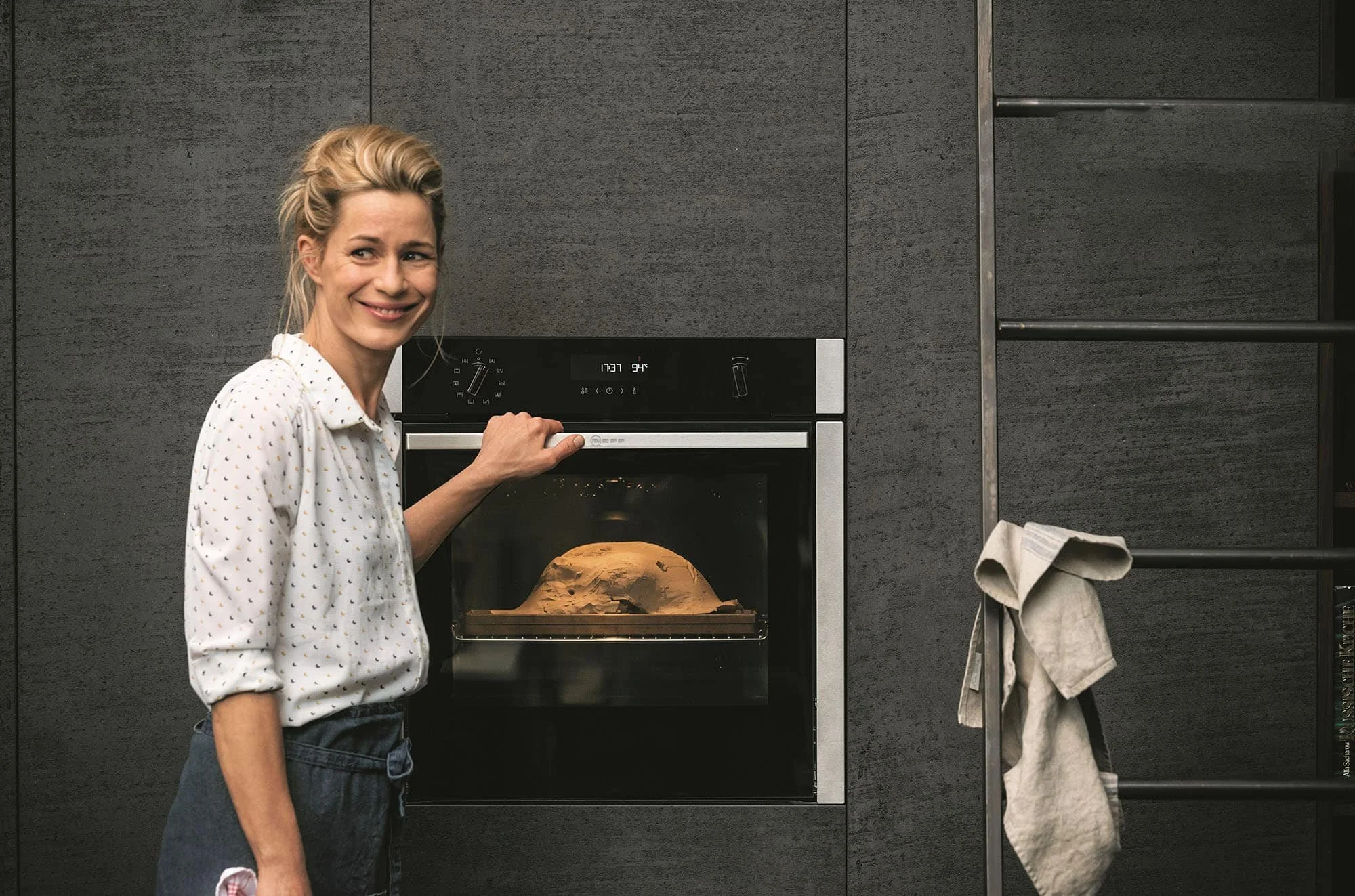 A woman with blonde hair smiling while opening a modern oven with a baked item inside in a kitchen with dark walls.