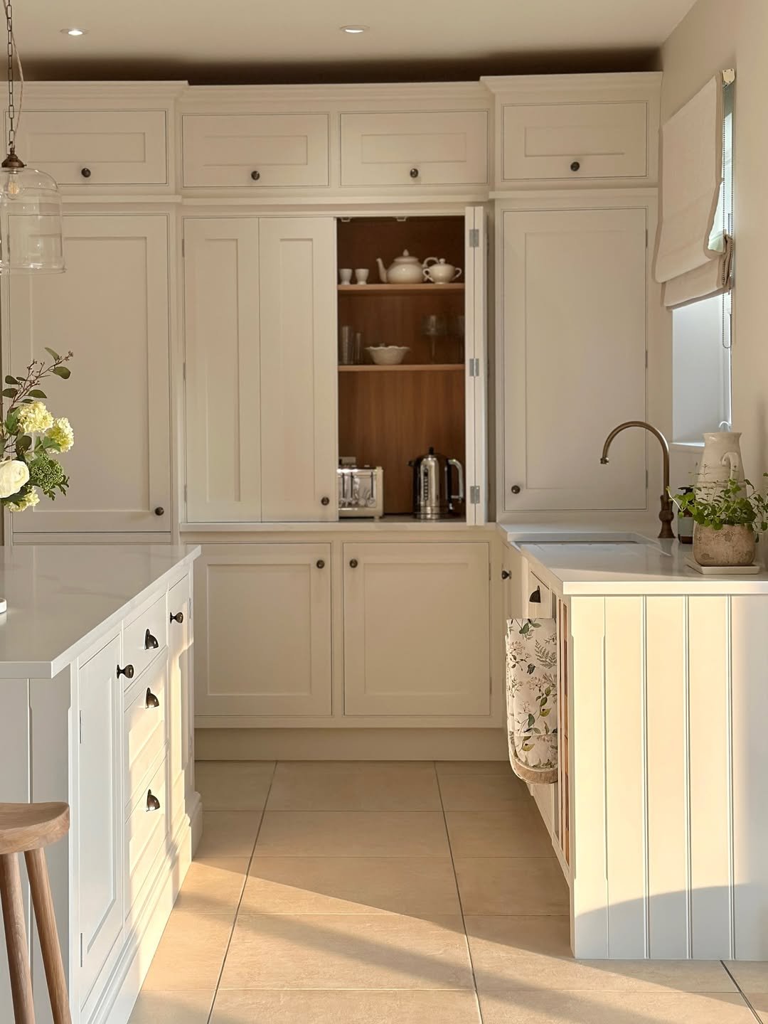 Bright kitchen with white cabinets, a small sink, and a window with a Roman shade. Open shelves with white dishes and small appliances, sunlight casting shadows on the tiled floor.