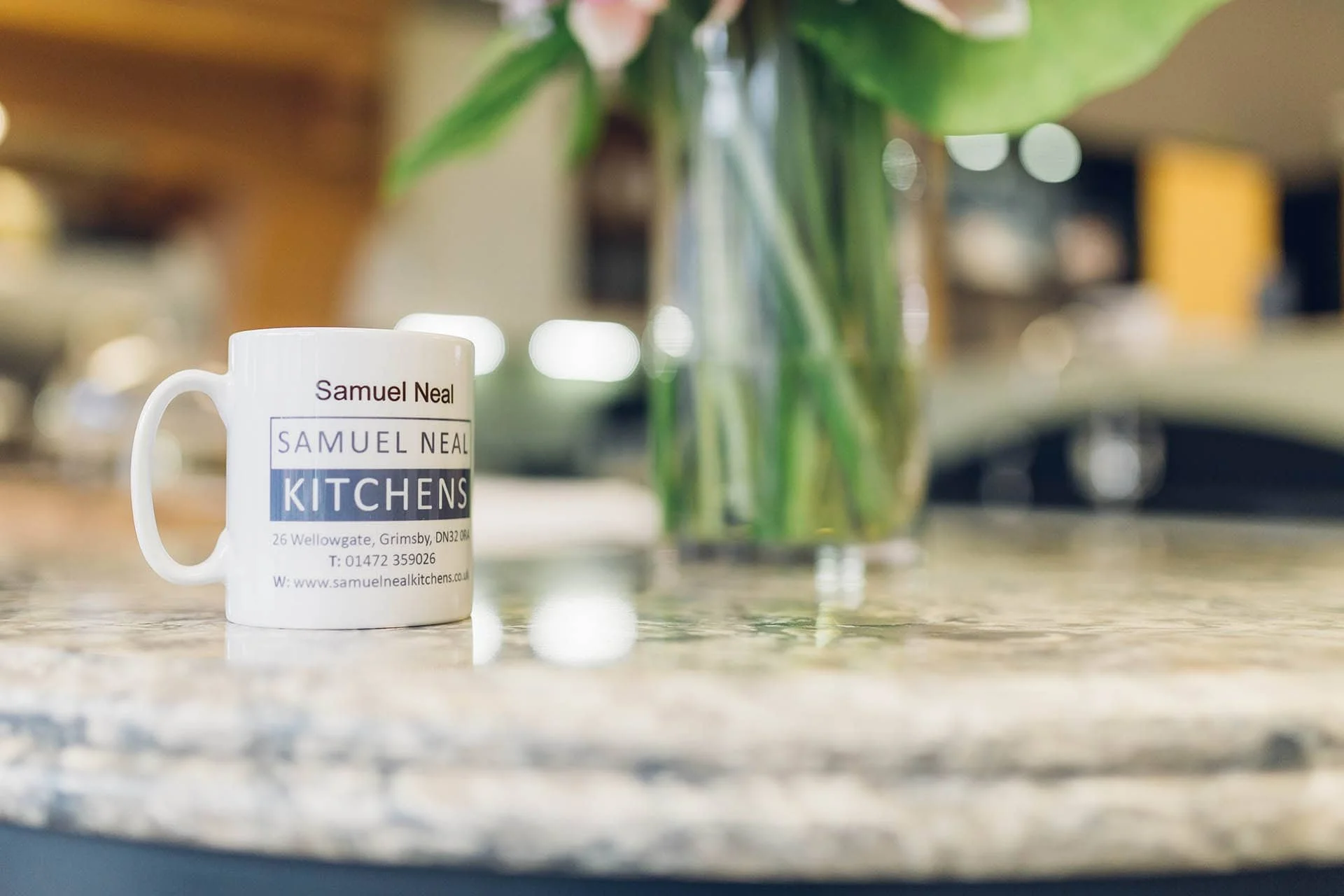 A white coffee mug with the text 'Samuel Neal KITCHENS' on it, placed on a marble countertop, with a blurred vase of flowers in the background.