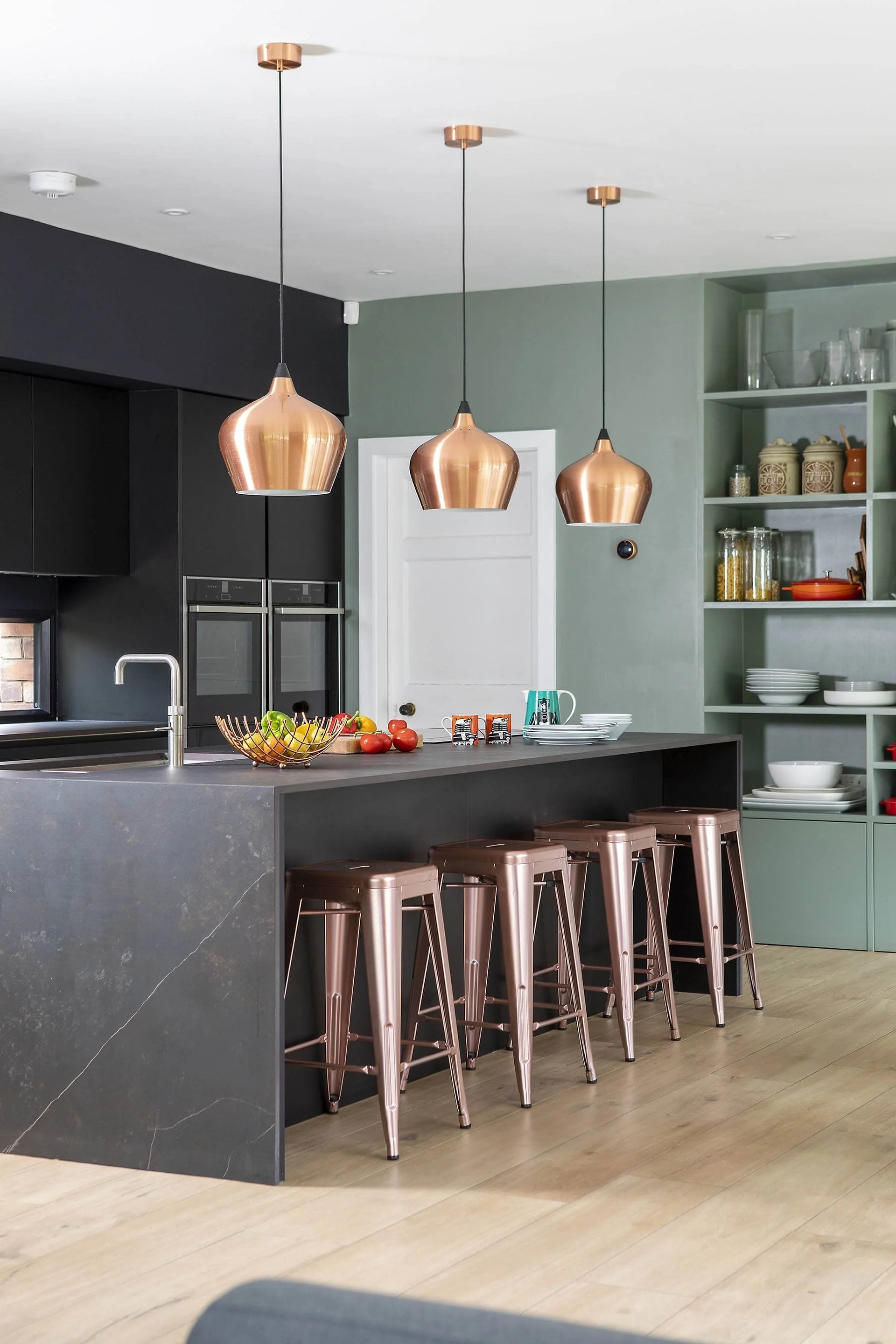Modern kitchen with black cabinets, green open shelving, copper pendant lights, a gray island with bar stools, and various kitchenware and fresh produce on the island.