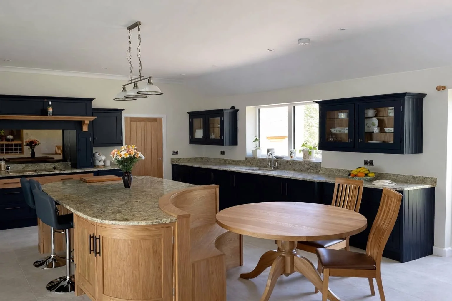 Kitchen with light wooden and dark navy cabinets, granite countertops, a kitchen island with a vase of flowers, and a round wooden dining table with chairs, illuminated by natural light from a window.