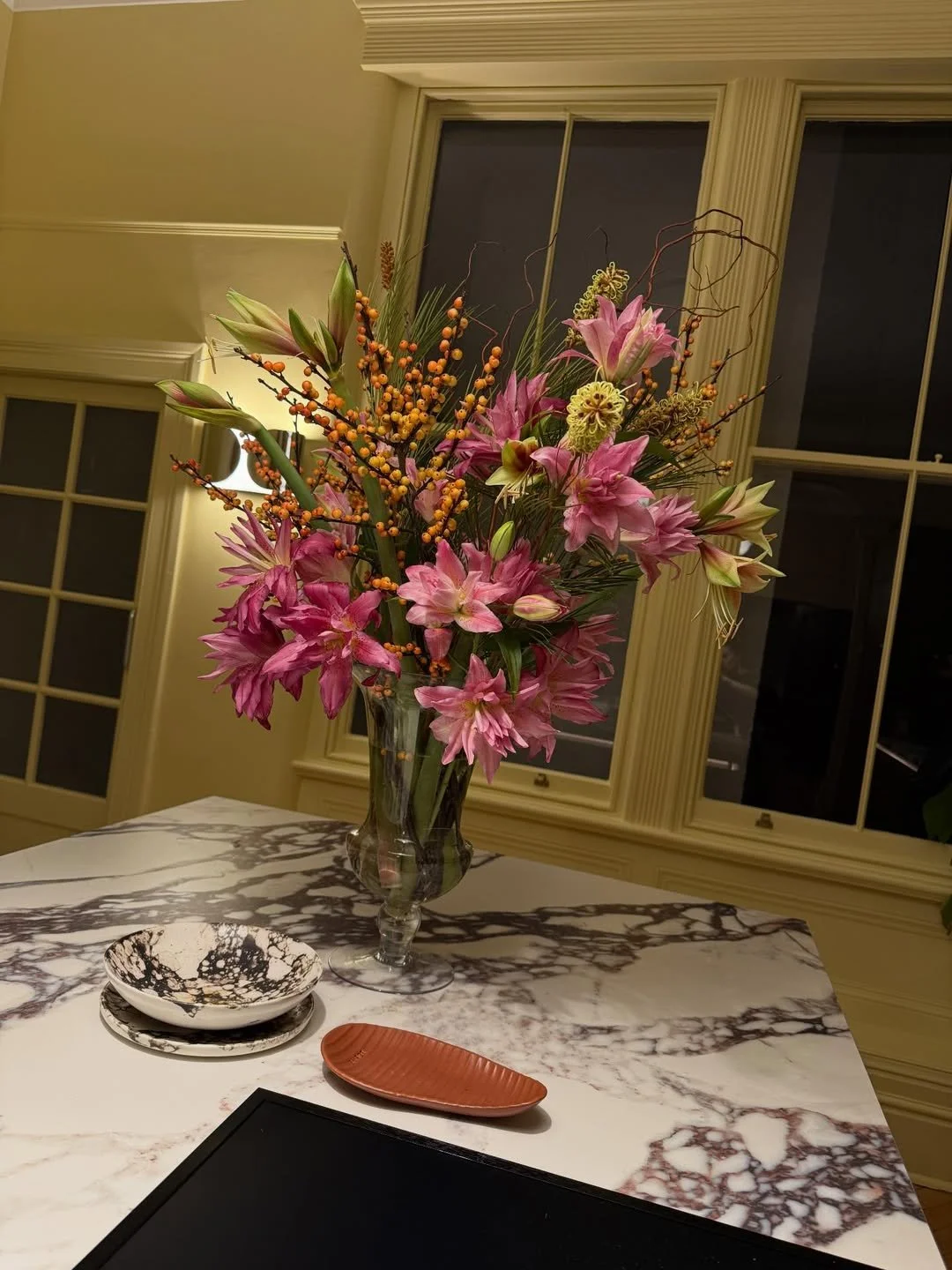 A floral arrangement featuring pink lilies, pink amaryllis, and orange berries in a clear glass vase on a marble table inside a room with large windows.