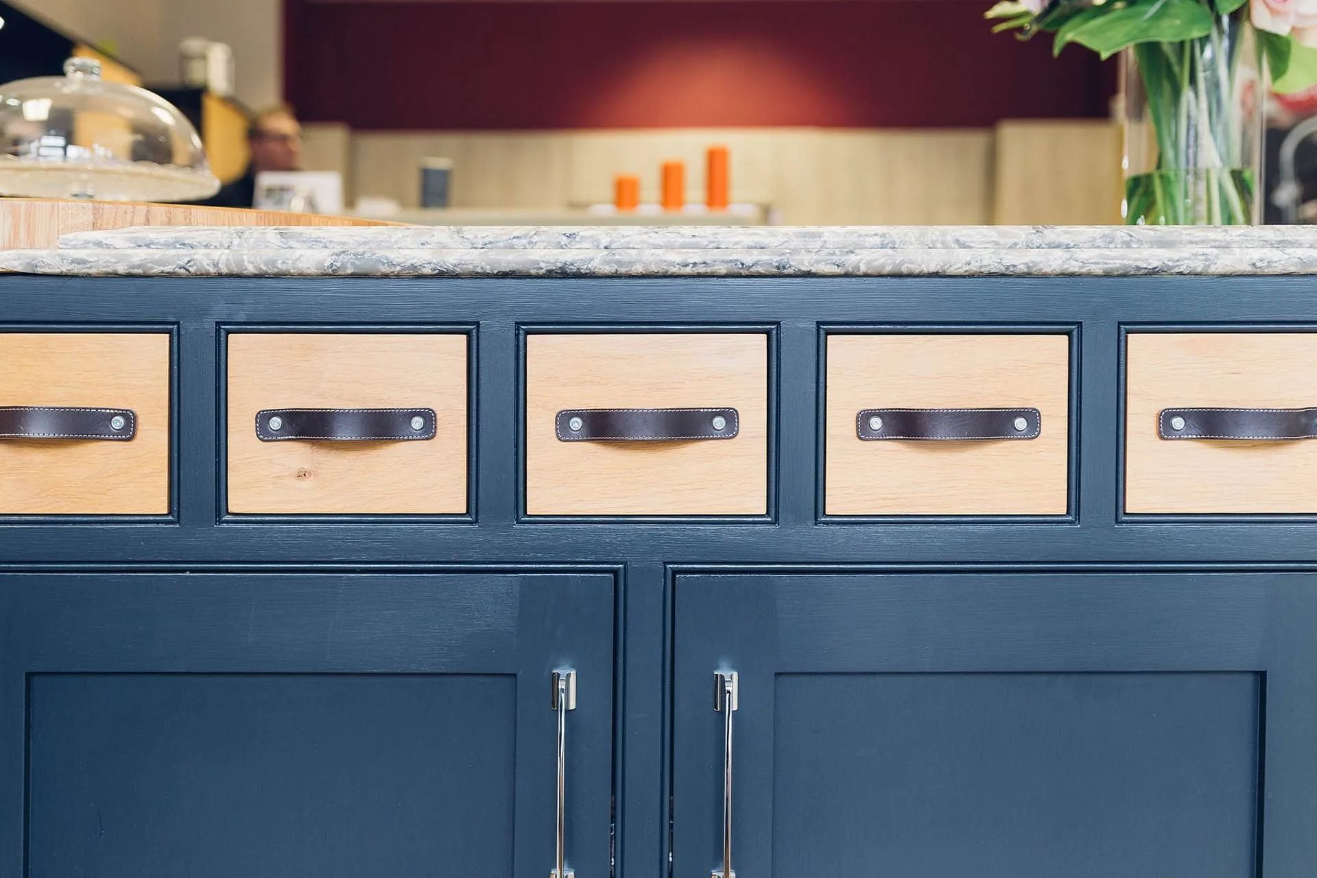 Close-up of a navy blue cabinet with multiple drawers and black leather handles topped with a gray marble countertop. In the background, there is a blurred café setting with a person and decoration.