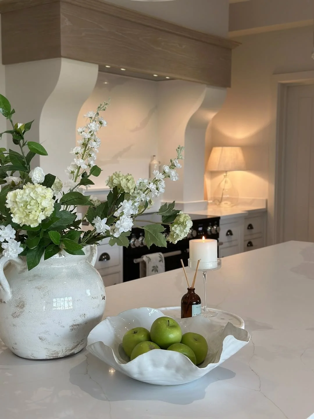 A white vase with white and green flowers, a white bowl with green apples, a lit candle, and diffuser sticks on a white kitchen countertop with a beige lamp and a black stove in the background.