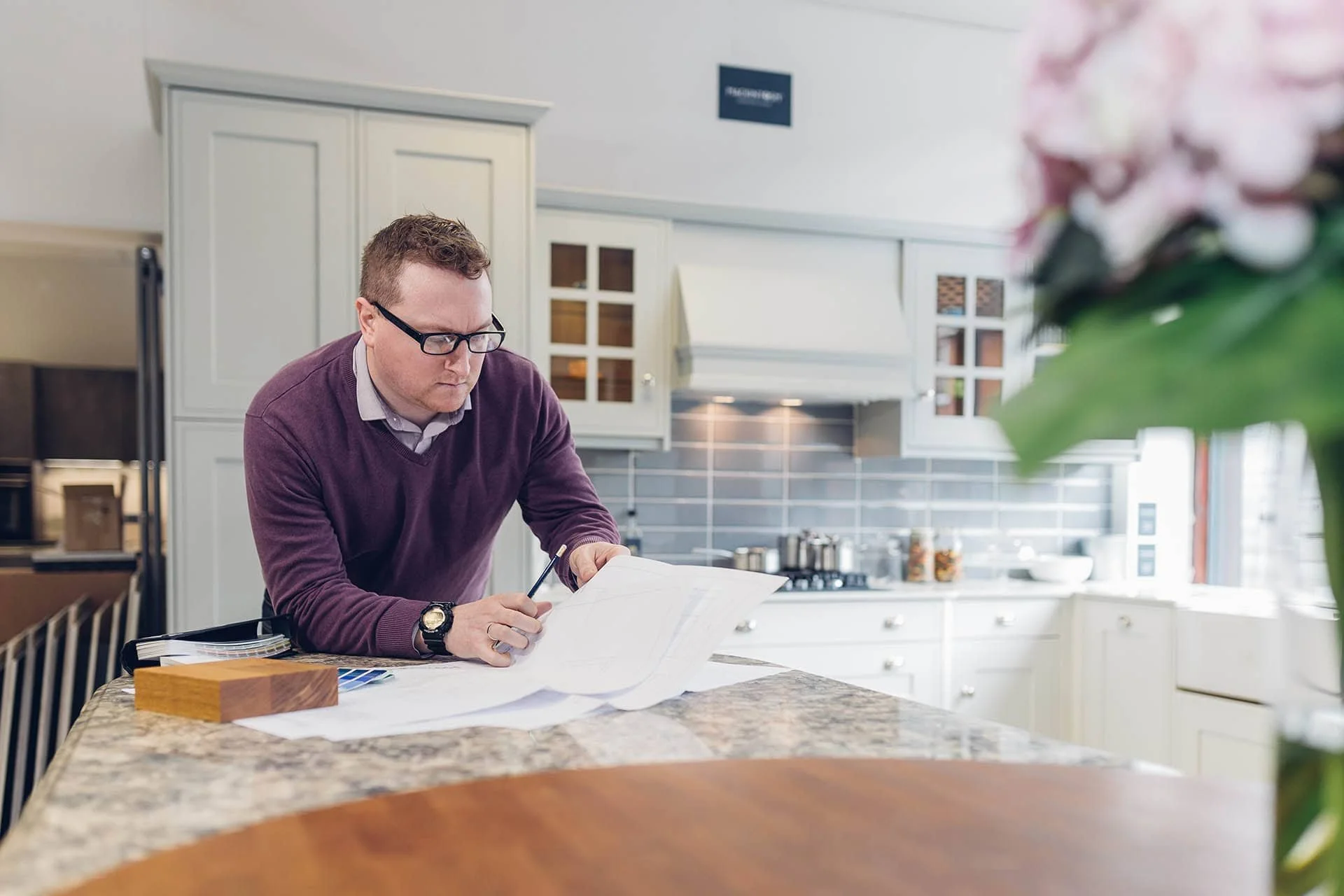 Man with glasses and a purple sweater working with blueprints at a kitchen island.