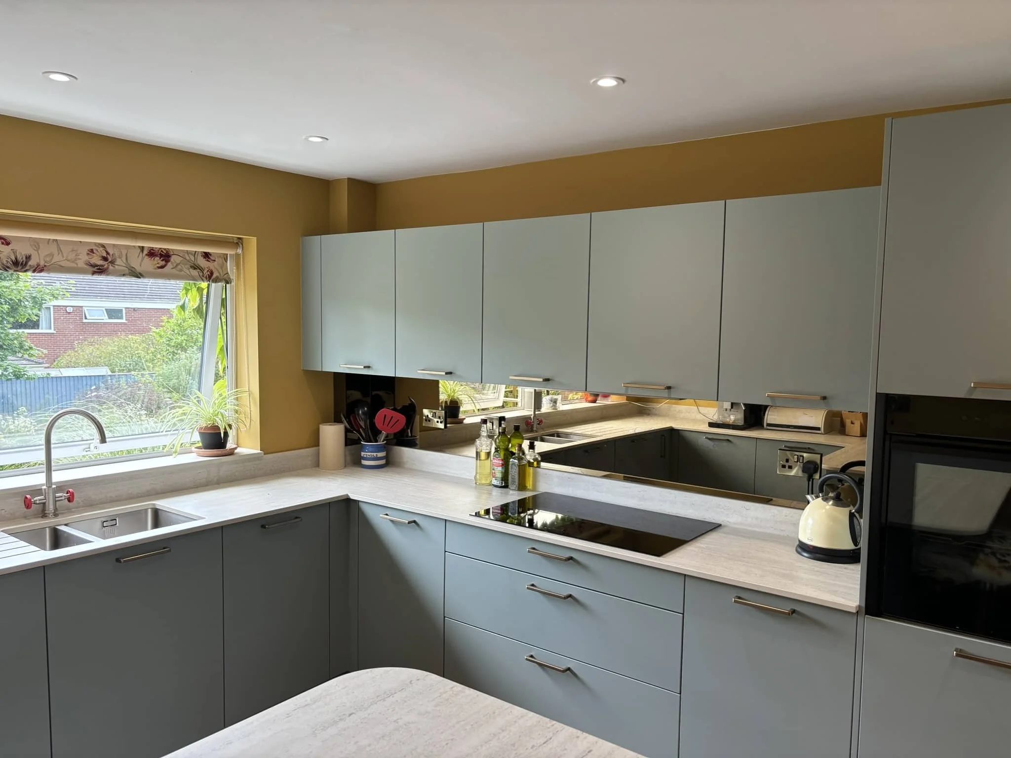Modern kitchen with gray cabinets, white countertops, a window with a view of trees and houses, and various small appliances and utensils on the counter.