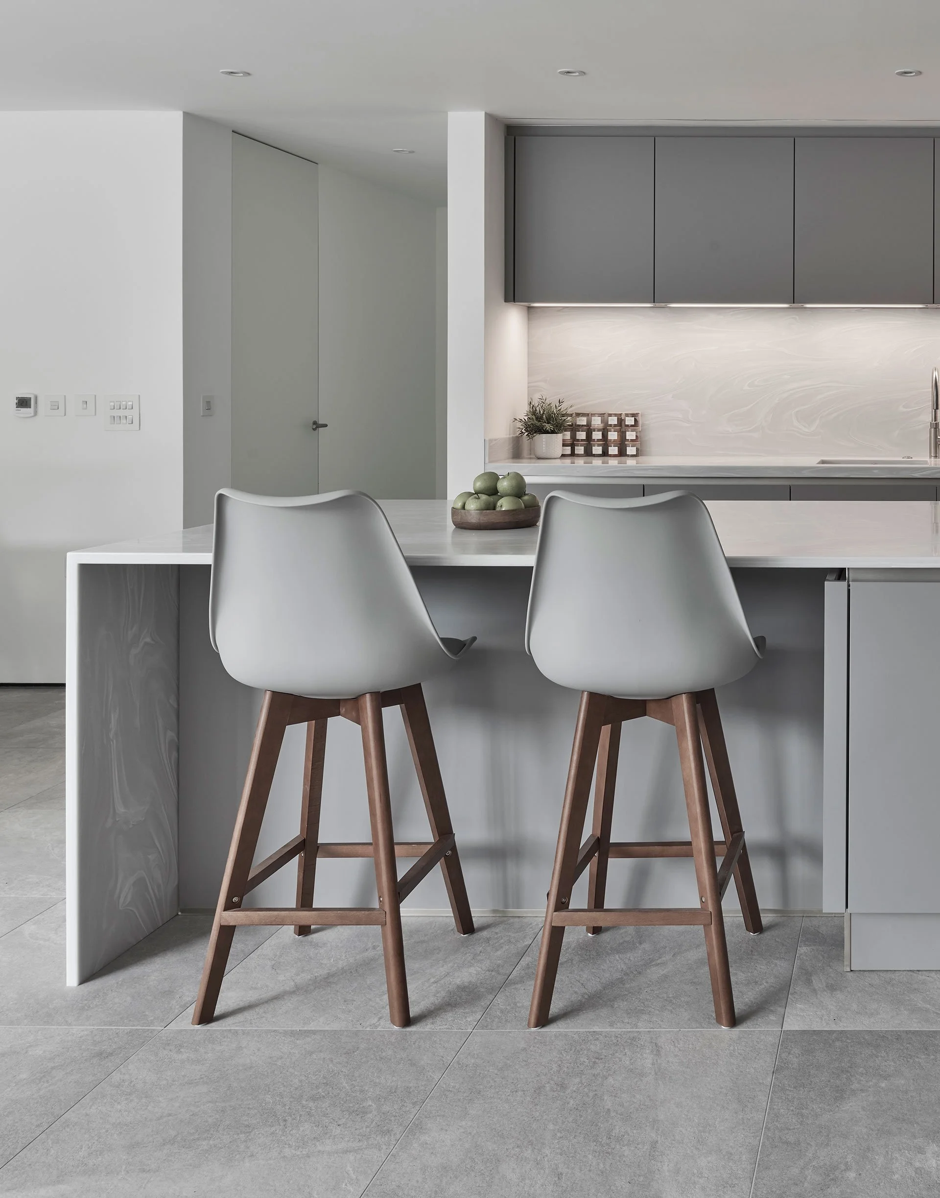 Modern kitchen with a white island counter and two gray bar stools with wooden legs. Gray cabinets and a marble backsplash are visible in the background.