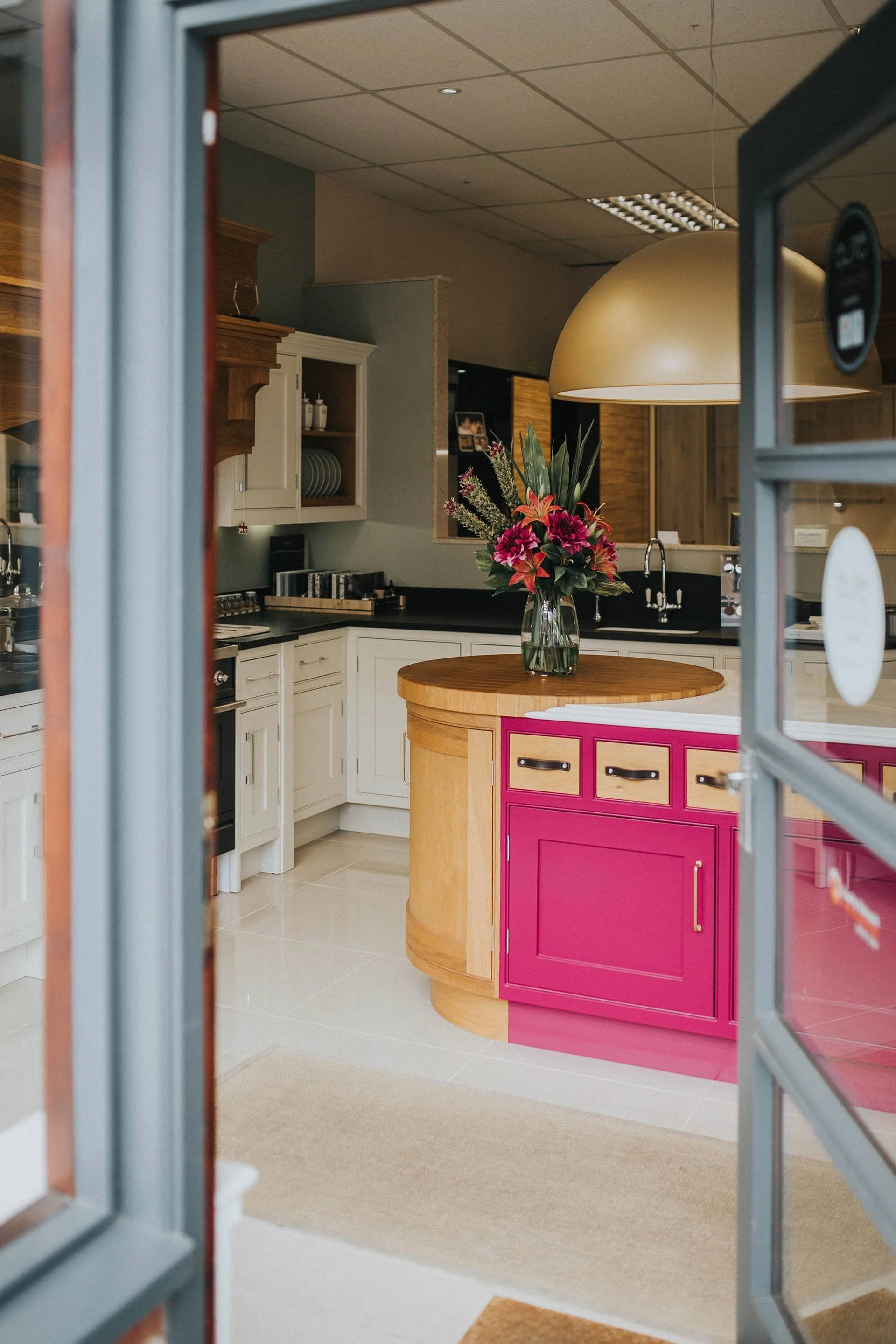 Kitchen seen through partially open glass door with pink cabinet and flower vase on wooden island.