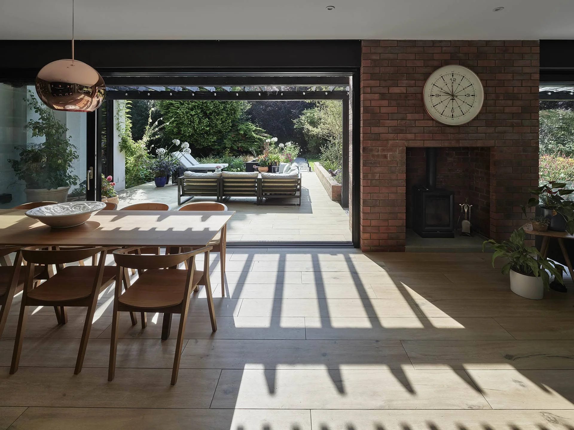 Interior view of a dining area with wooden table and chairs, adjacent to a brick fireplace with a clock, opening to a patio with outdoor seating and garden.