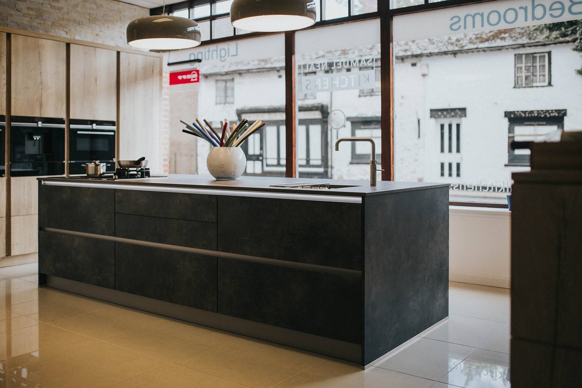 Modern kitchen with a dark island counter, a white bowl with colorful utensils, a sink, and large windows showing a building outside.