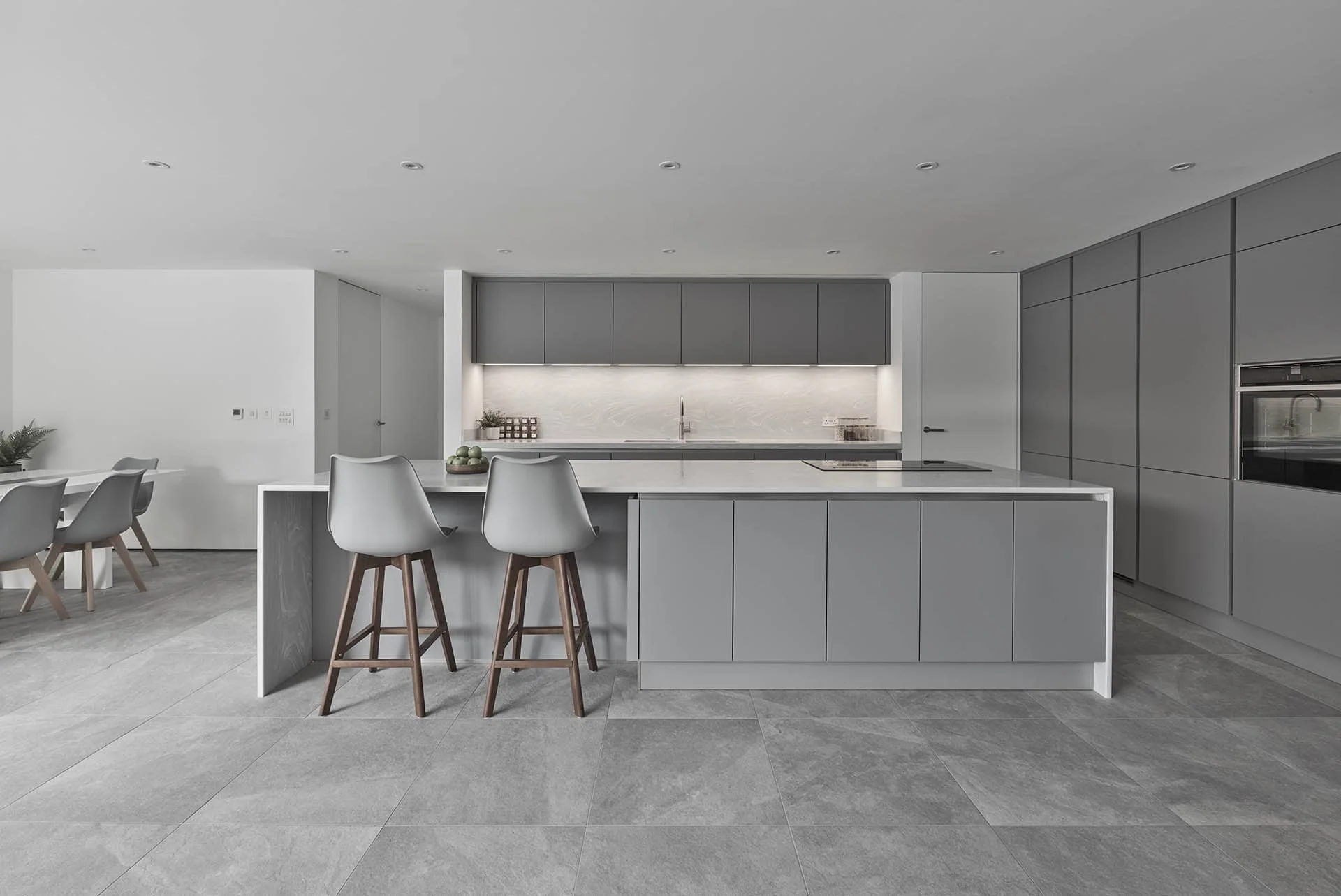 Modern minimalist kitchen with gray cabinets, white countertops, and a large island with seating for three. The floor is tiled in gray. There is a dining area visible to the left with a table and gray chairs.