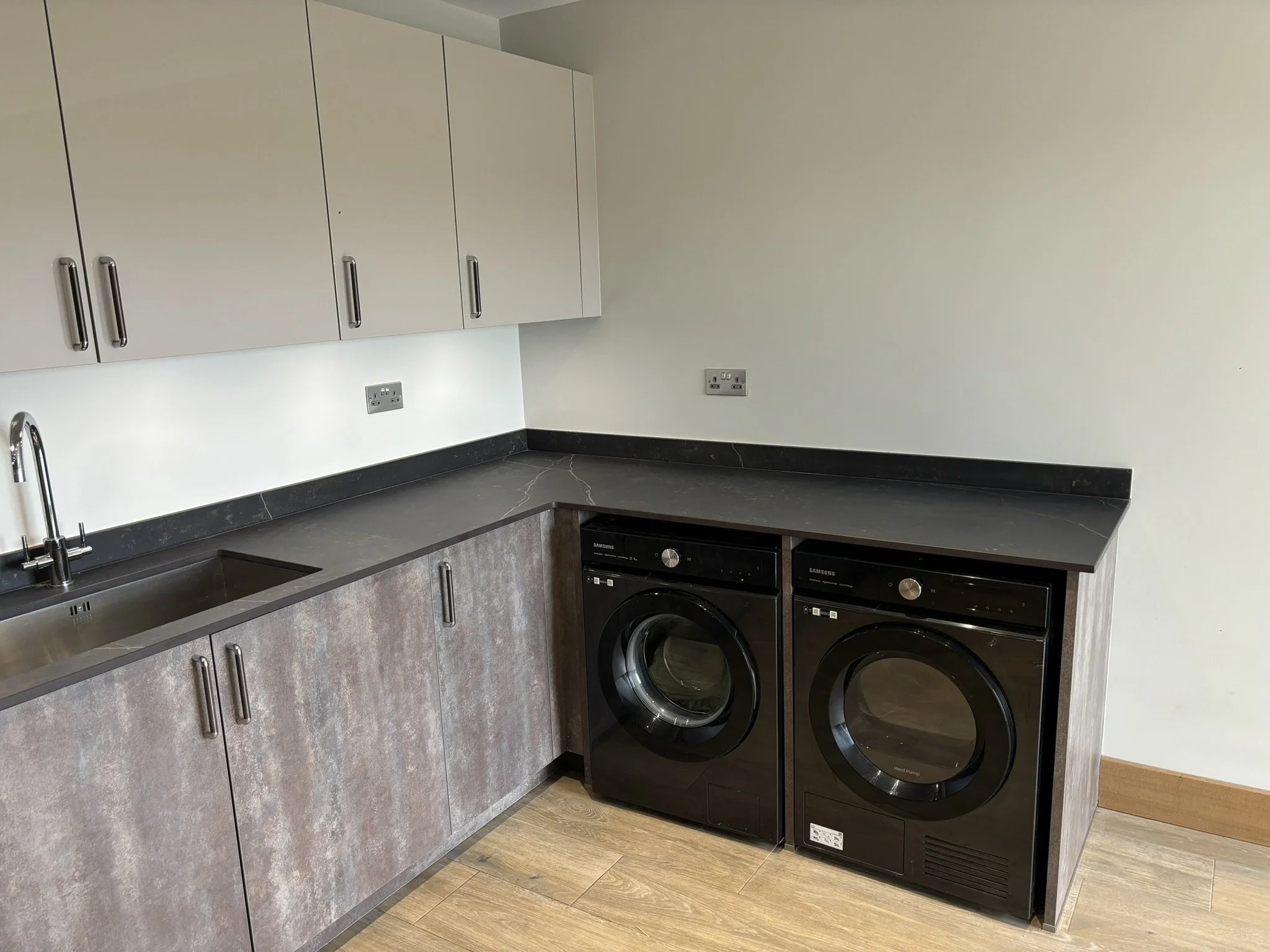 A laundry room with a black washing machine and a black dryer side by side under a black countertop. The room has white upper cabinets with silver handles, a greyish lower cabinet, gray backsplash, and light wood flooring.
