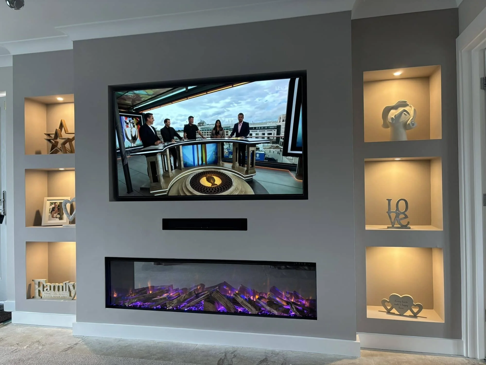 Living room wall with a mounted TV showing a TV studio and an electric fireplace below it. Recessed shelves with decorative items, including a star, a framed photo, a heart, a 'LOVE' sign, and a sign reading 'Family'.