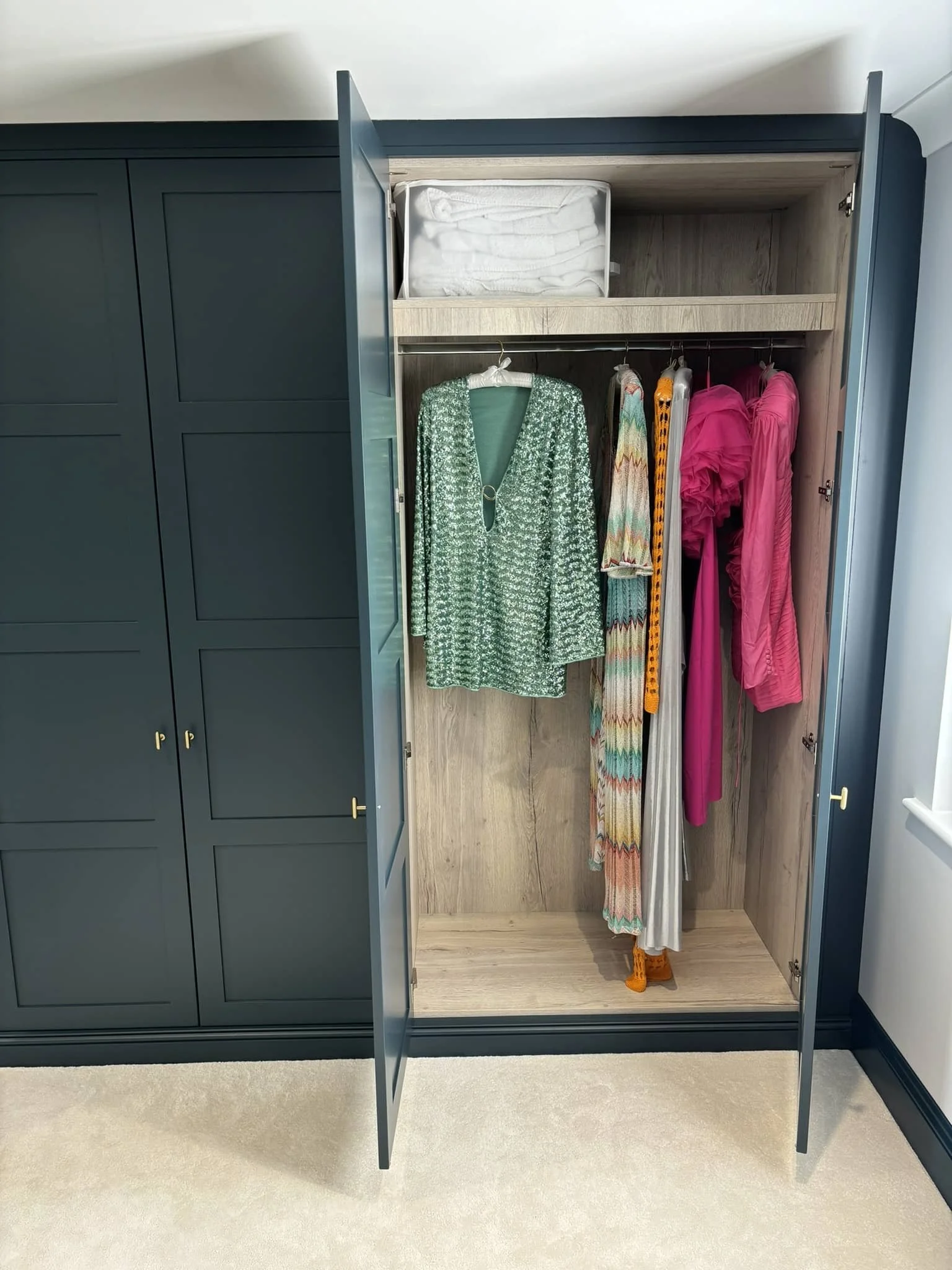 Open wardrobe with hanging clothes, including a green patterned top, colorful dress, pink dresses, and a pair of orange shoes underneath.