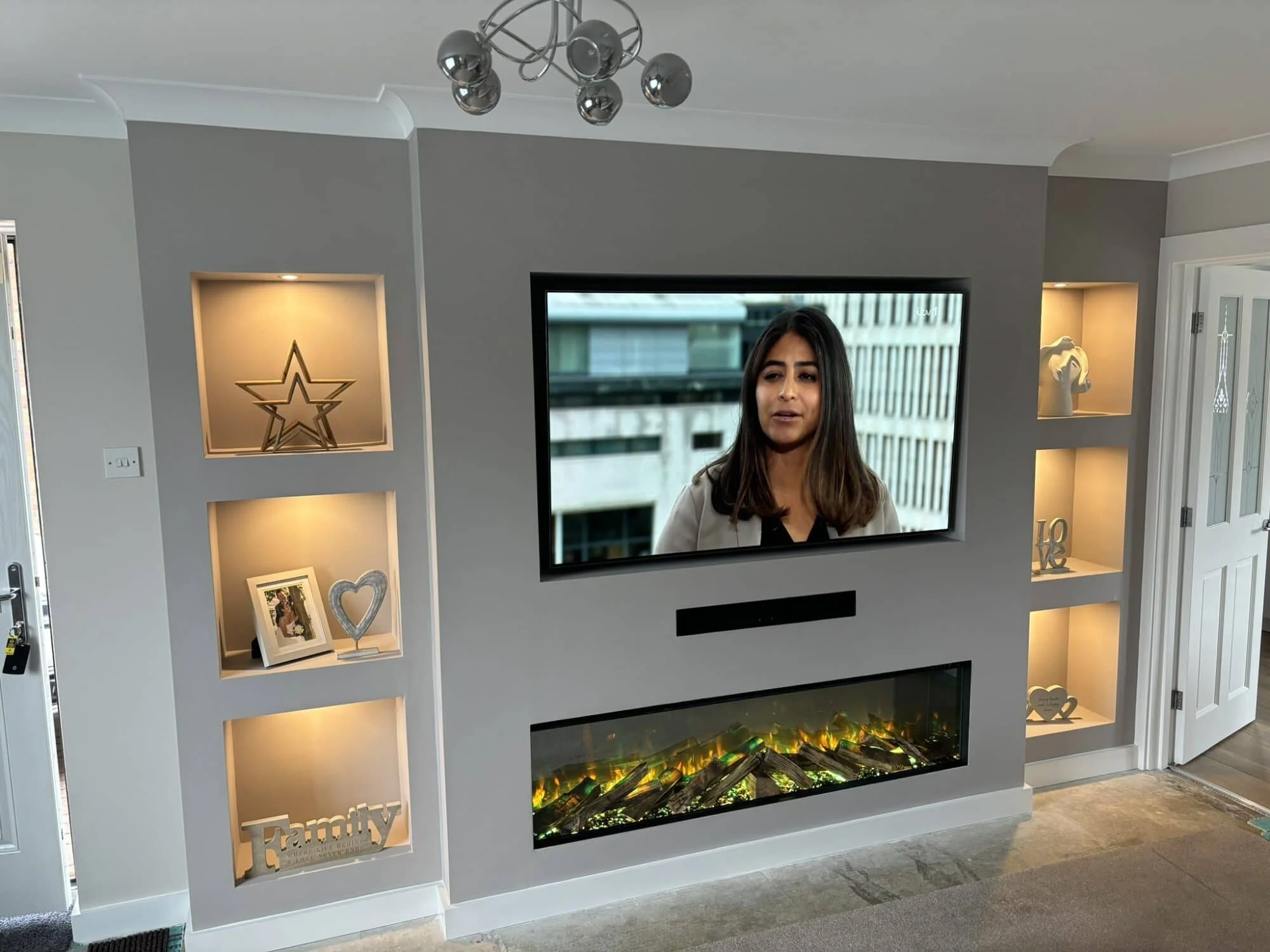 Living room with a wall-mounted TV showing a woman, decorative shelves with lights and ornaments, electric fireplace below the TV, and a chandelier overhead.