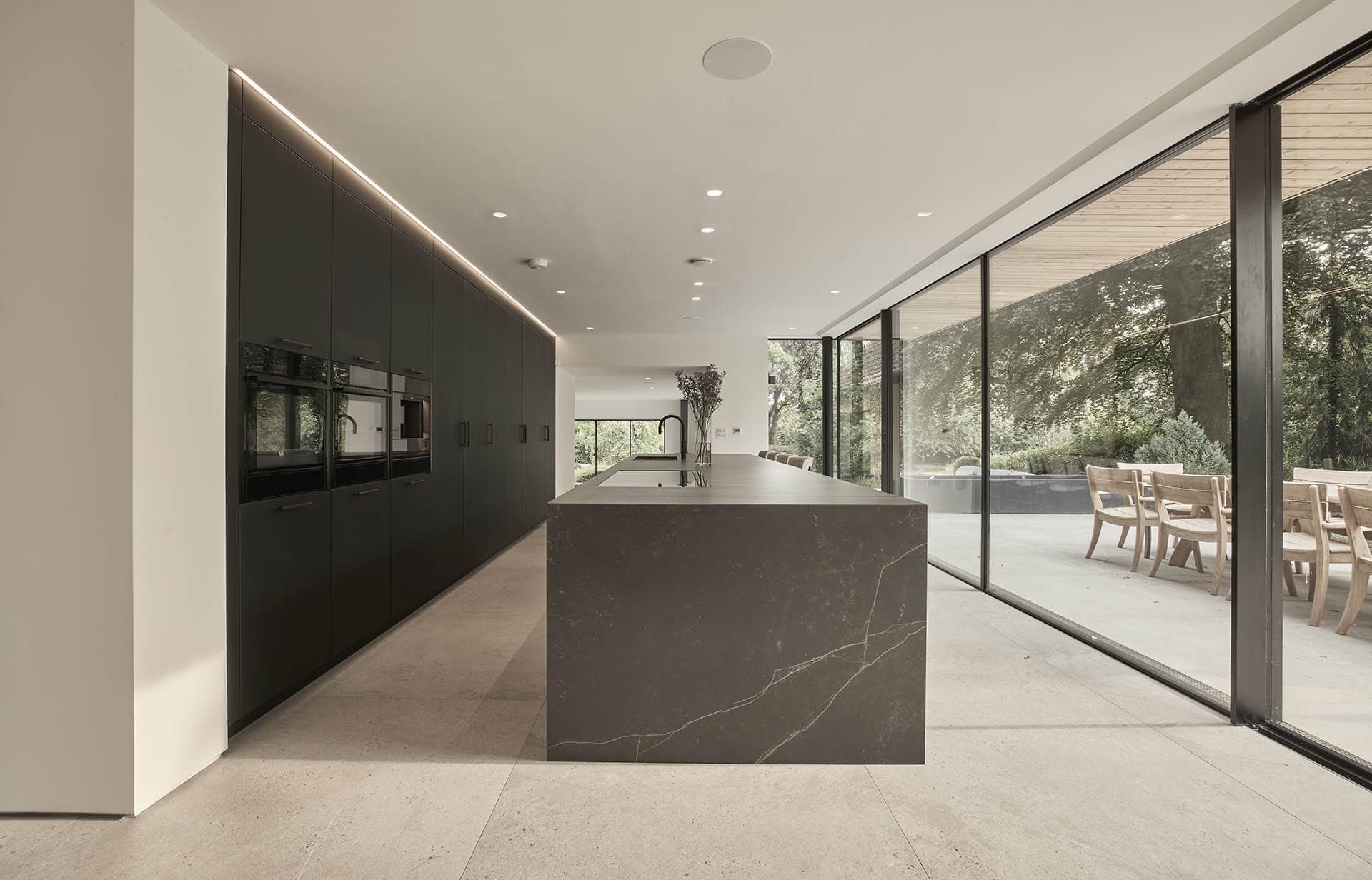 Modern kitchen with black cabinetry, large marble island, and floor-to-ceiling glass doors leading to outdoor seating area.