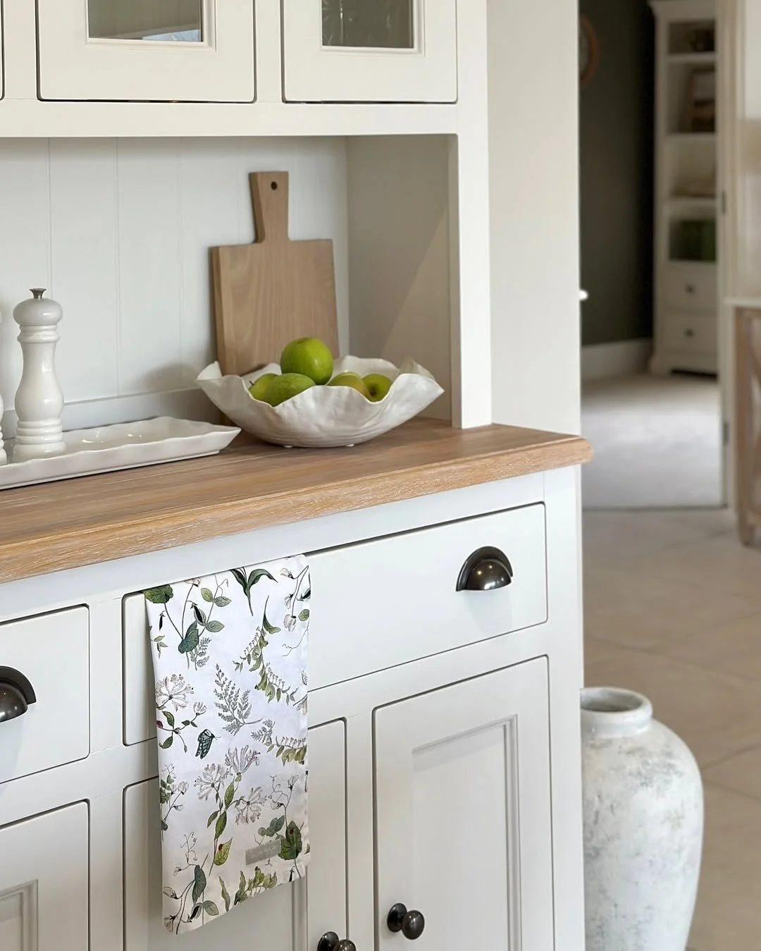 A kitchen counter with a white ceramic bowl filled with green apples, a wooden cutting board, a white ceramic tray, and a tablecloth with a fern and leaf design hanging from a cabinet. There is also a large white vase on the floor to the right.