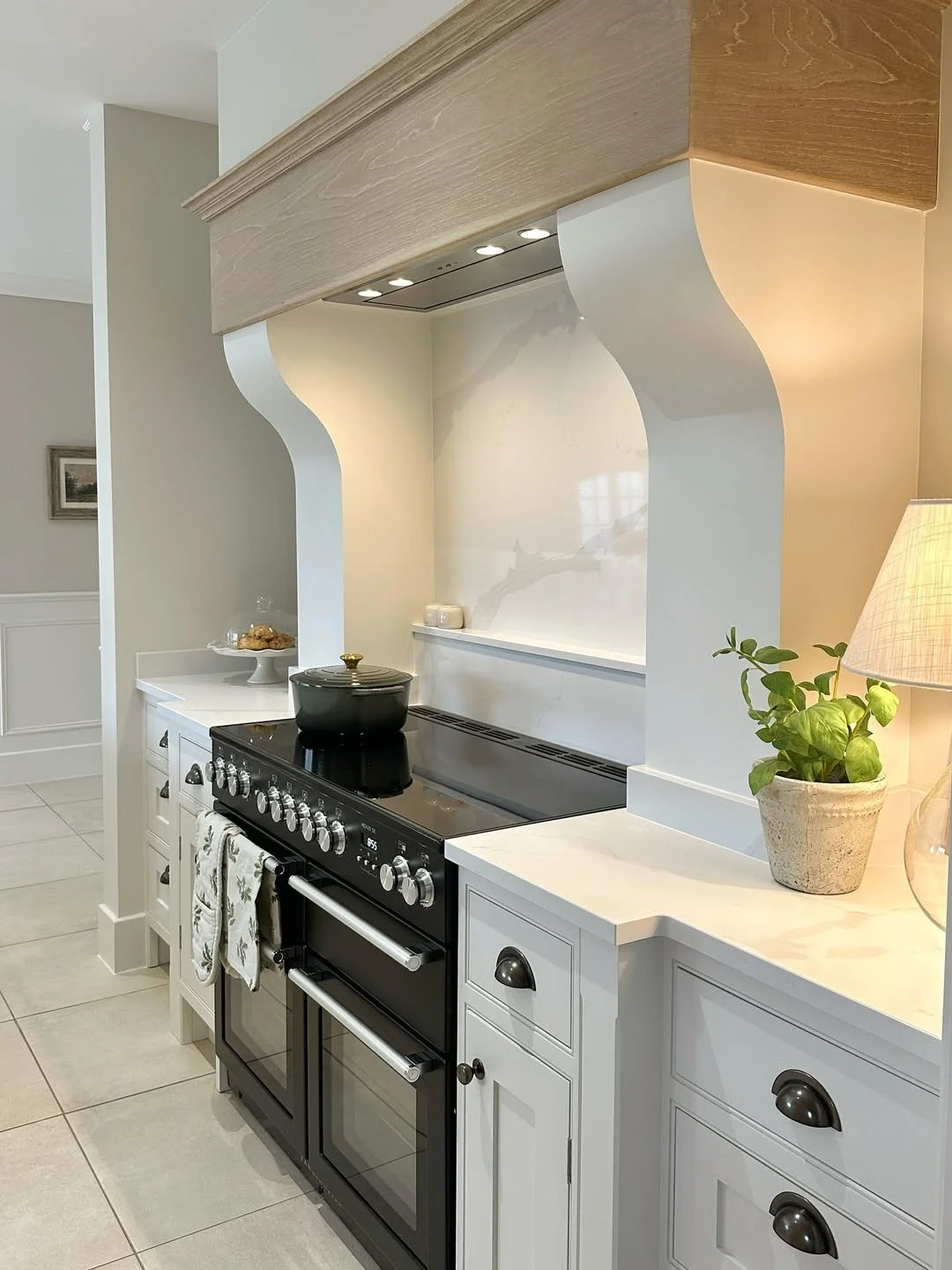 Kitchen with black stove, white cabinets, potted plant, beige tiled floor, and wooden range hood.