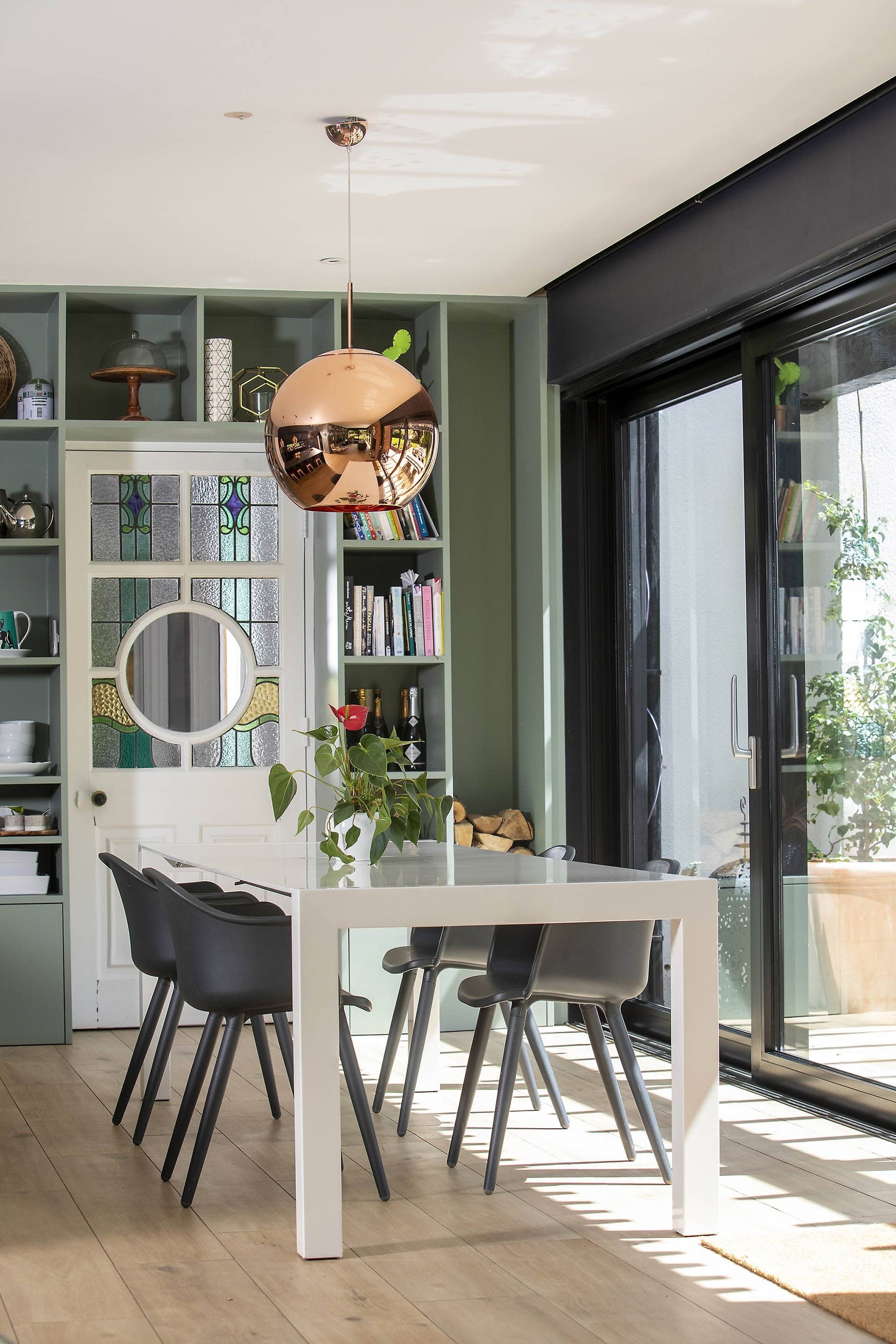 Modern dining room with a white table, black and gray chairs, a potted plant, green shelves with books and decor, large sliding glass door, and a copper pendant light.