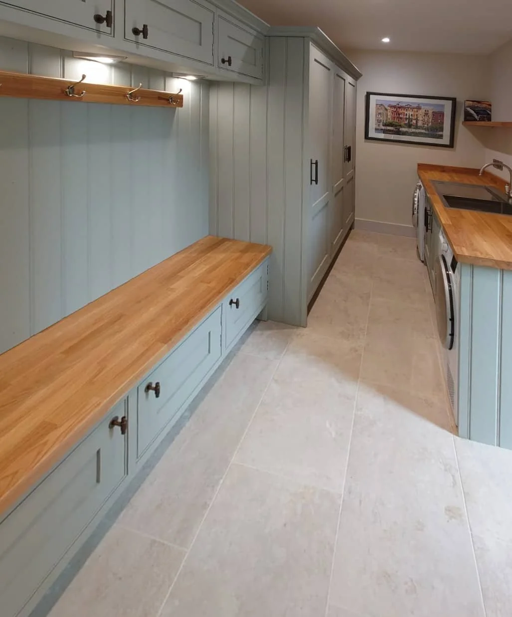 A kitchen with light blue cabinets and drawers, wooden countertops, a wall-mounted wooden coat rack with hooks, a framed picture hanging on the wall, and floor tiles.