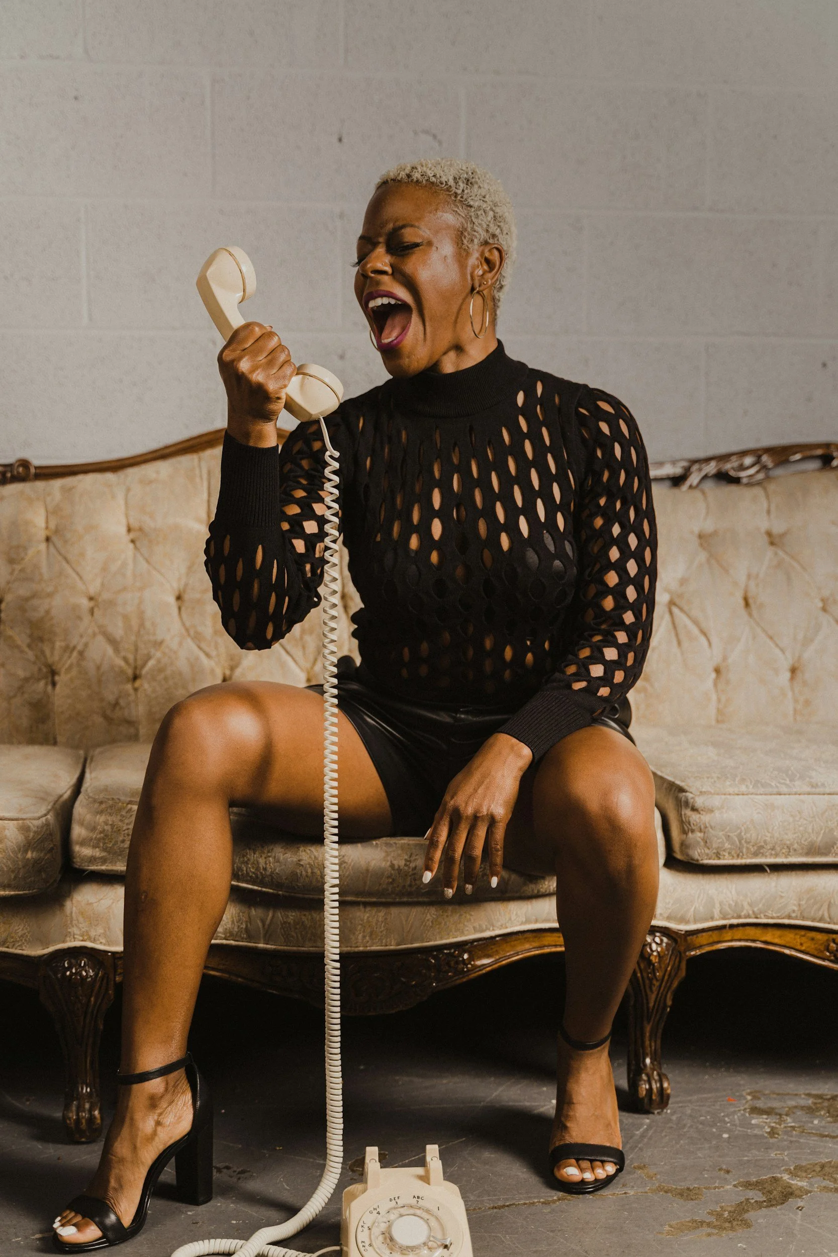 A woman with short blond hair and hoop earrings sitting on a vintage beige sofa, holding a rotary phone receiver to her mouth, with a lively expression as if she is shouting or singing.