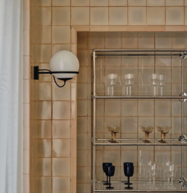 A beige tiled wall with a modern wall-mounted light fixture and a metal shelving unit holding three glasses and two black cups.