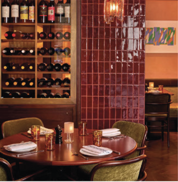 Interior of a restaurant with a round wooden table set for four, with napkins, glasses, and condiments. A wine rack with bottles is on the left, and a red tiled partition is in the center. There are green chairs around the table and some artwork on the wall in the background.