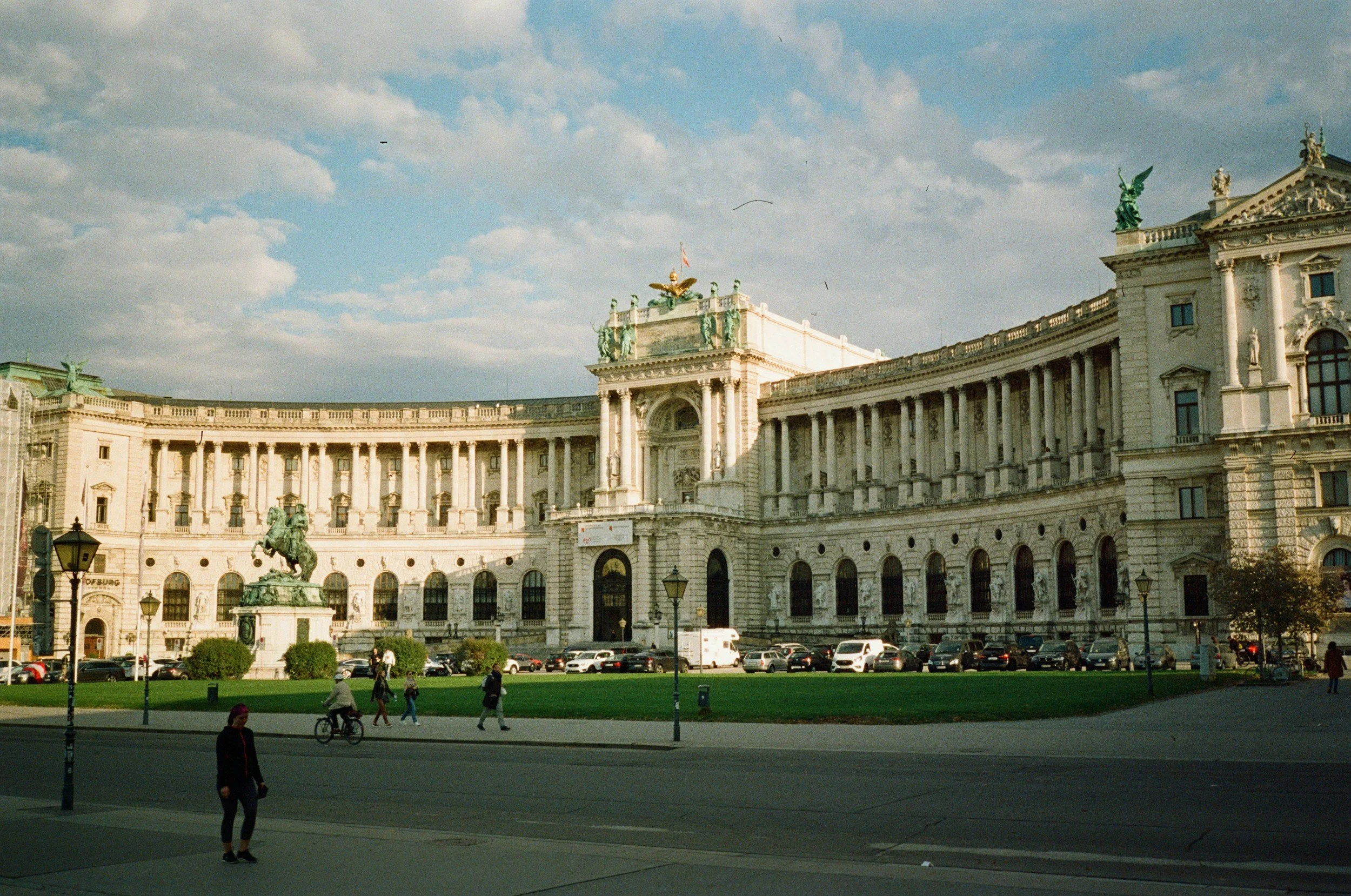 A historic building with classical architecture featuring arches, columns, and statues, situated on a city street with parked cars, pedestrians, and a statue of a rider on a horse in front.