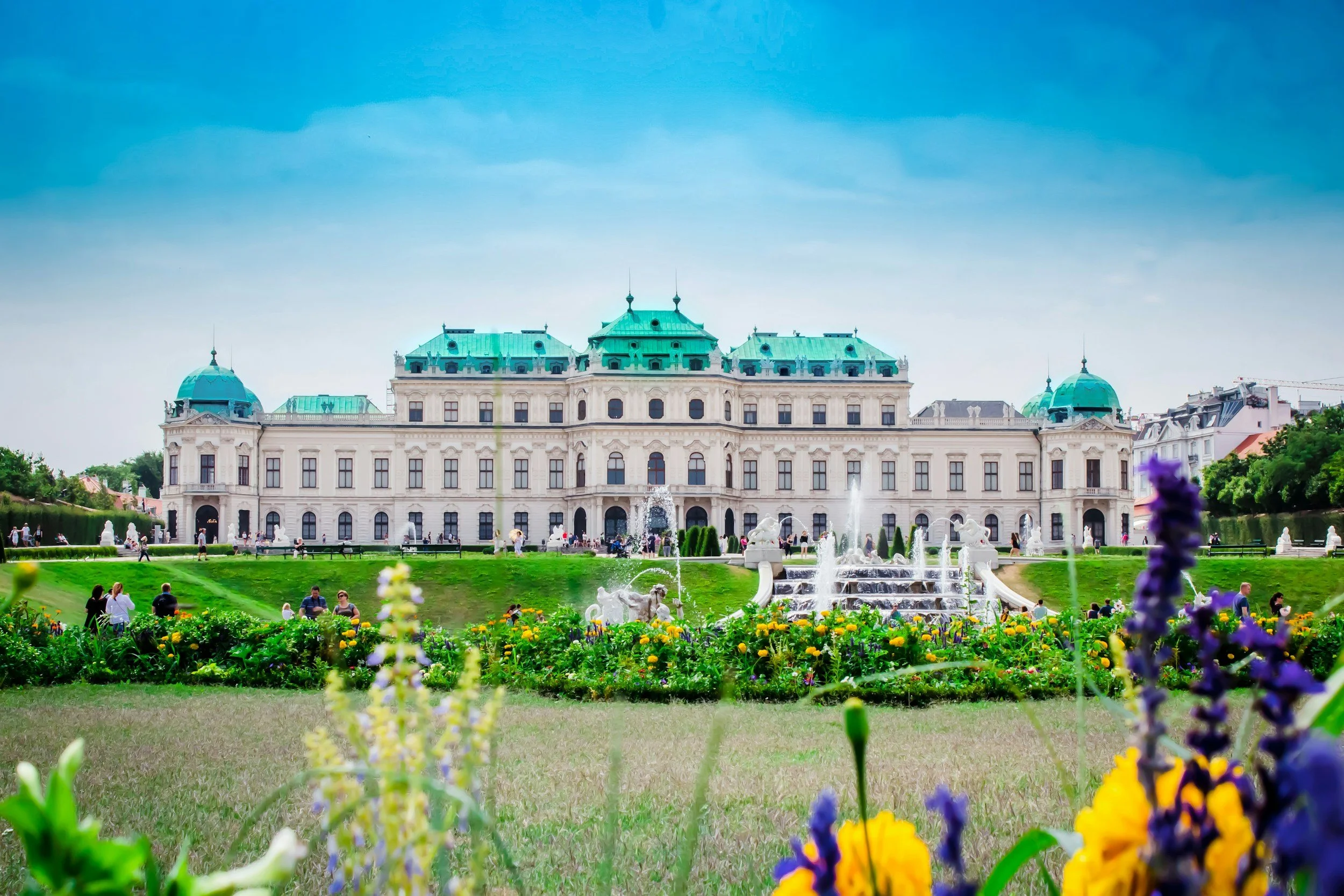 A large historic palace with green domed roofs, surrounded by a lush garden with colorful flowers and fountains, under a bright blue sky.
