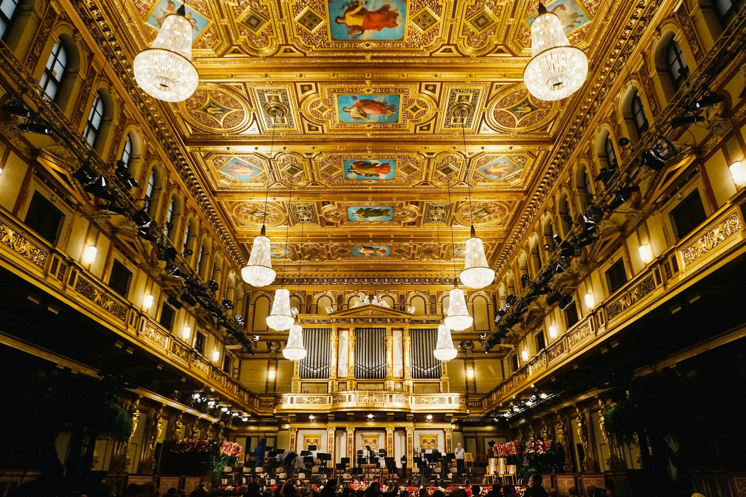 A grand concert hall with ornate golden decor, chandeliers hanging from an elaborately decorated ceiling featuring painted figures, and a stage with a group of musicians preparing for a performance.