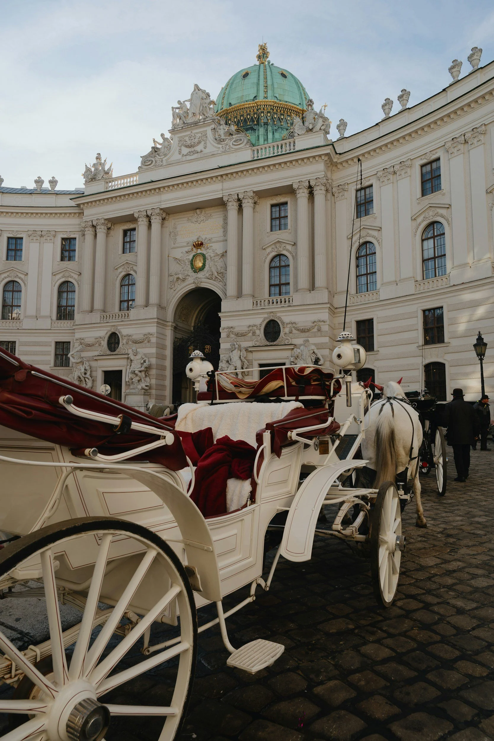 A white horse-drawn carriage with red and white blankets on its seats parked in front of a grand, historic building with columns, ornate sculptures, and a green dome in Vienna, Austria.