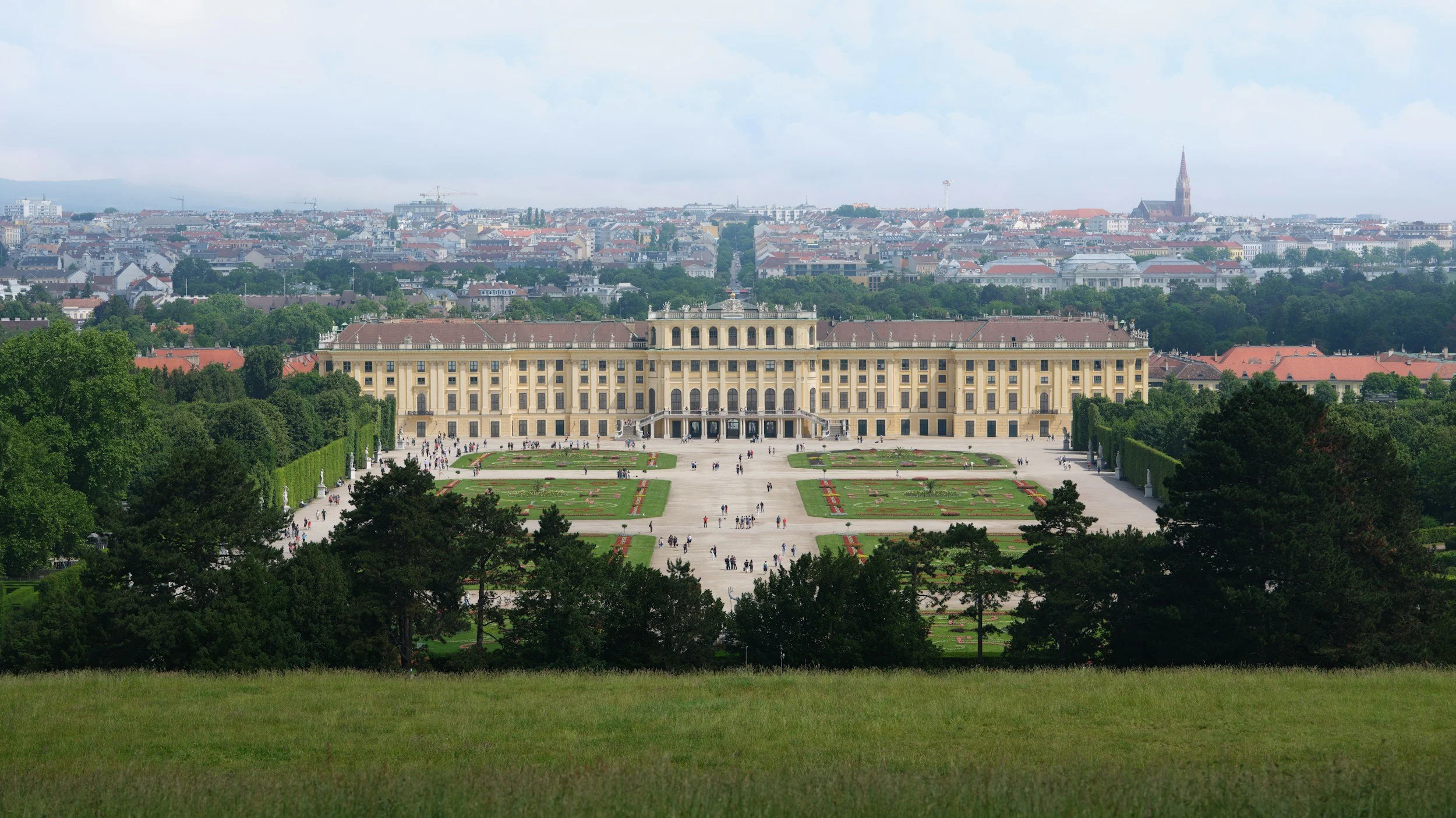 A grand historic palace with a large, manicured garden in front, surrounded by trees and a cityscape in the background. People are walking in the garden.